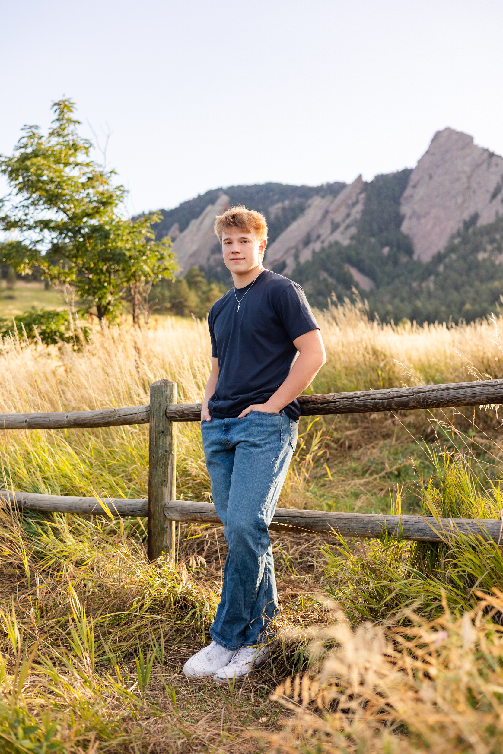 A young man leans against a fence at Chautauqua Park in Boulder, Colorado and smiles at the camera.