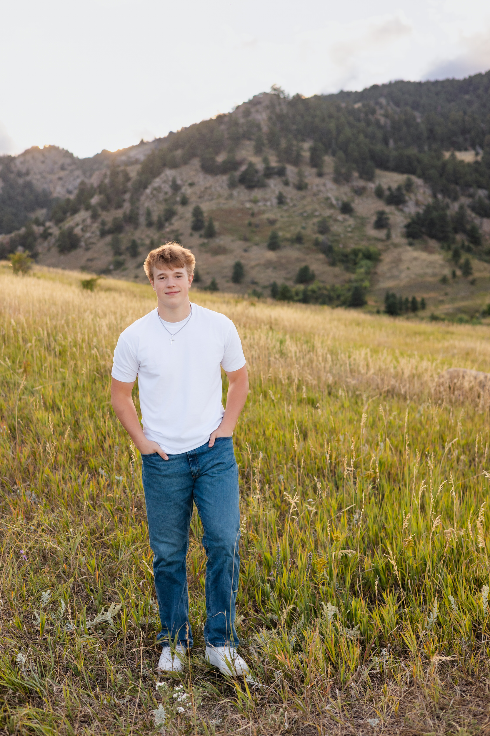 A young man stands in a field of tall grass with his hands in his pockets and smiles at the camera.