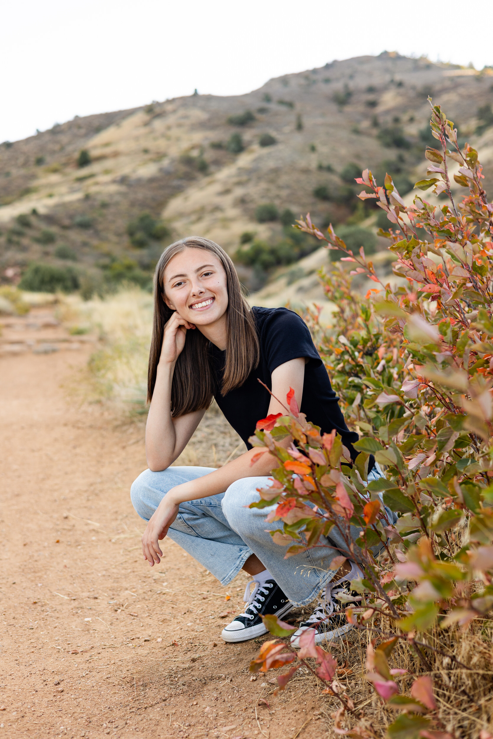 A young woman squats down on a dirt path and rests her chin on her fist and smiles at the camera.