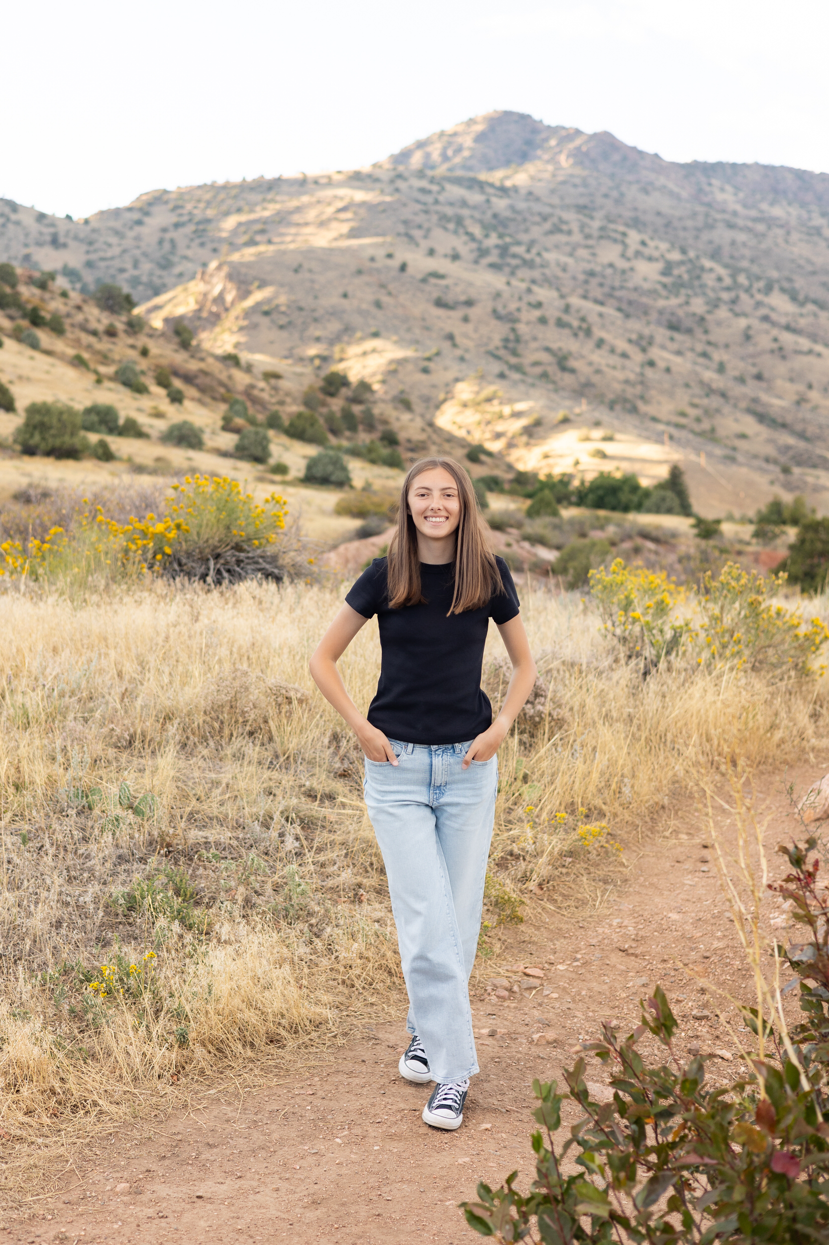 A young woman walks down a dirt path surrounded by tall grass with mountains in the background and smiles at the camera.