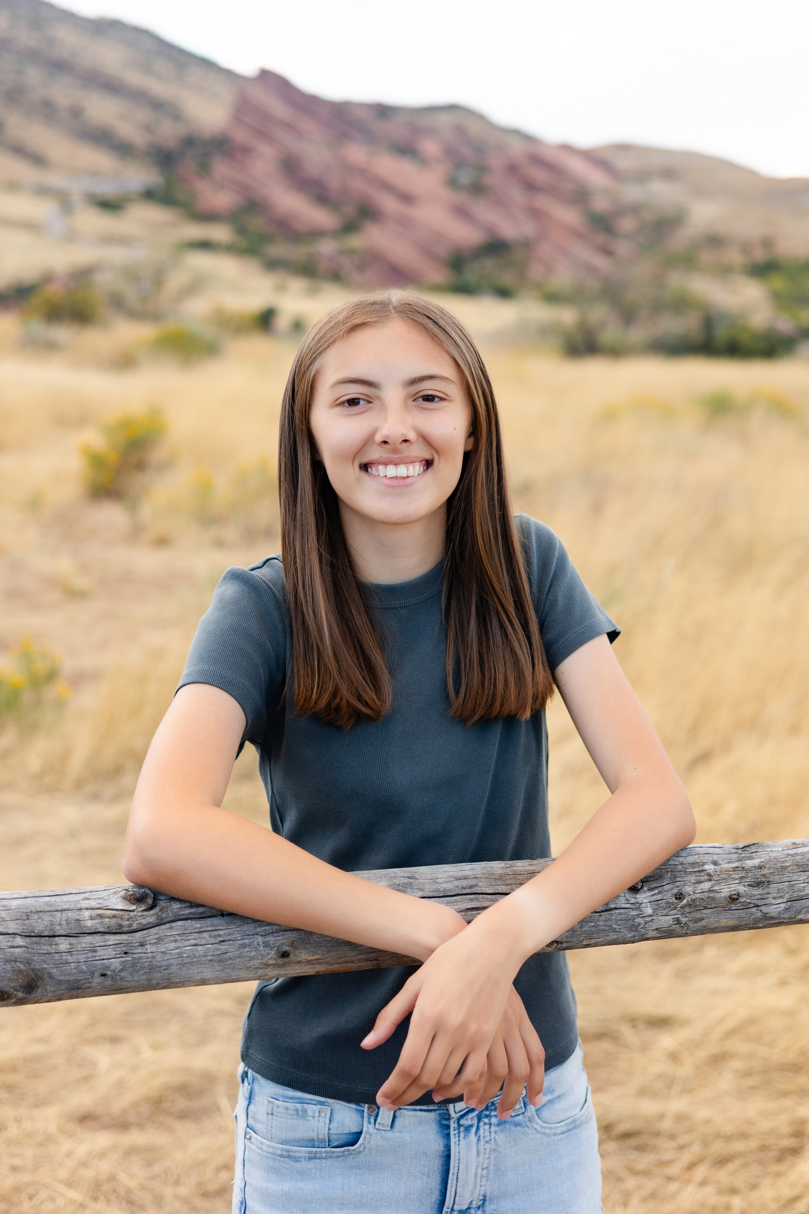 A young woman rests her forearms on a wooden fence and smiles at the camera.