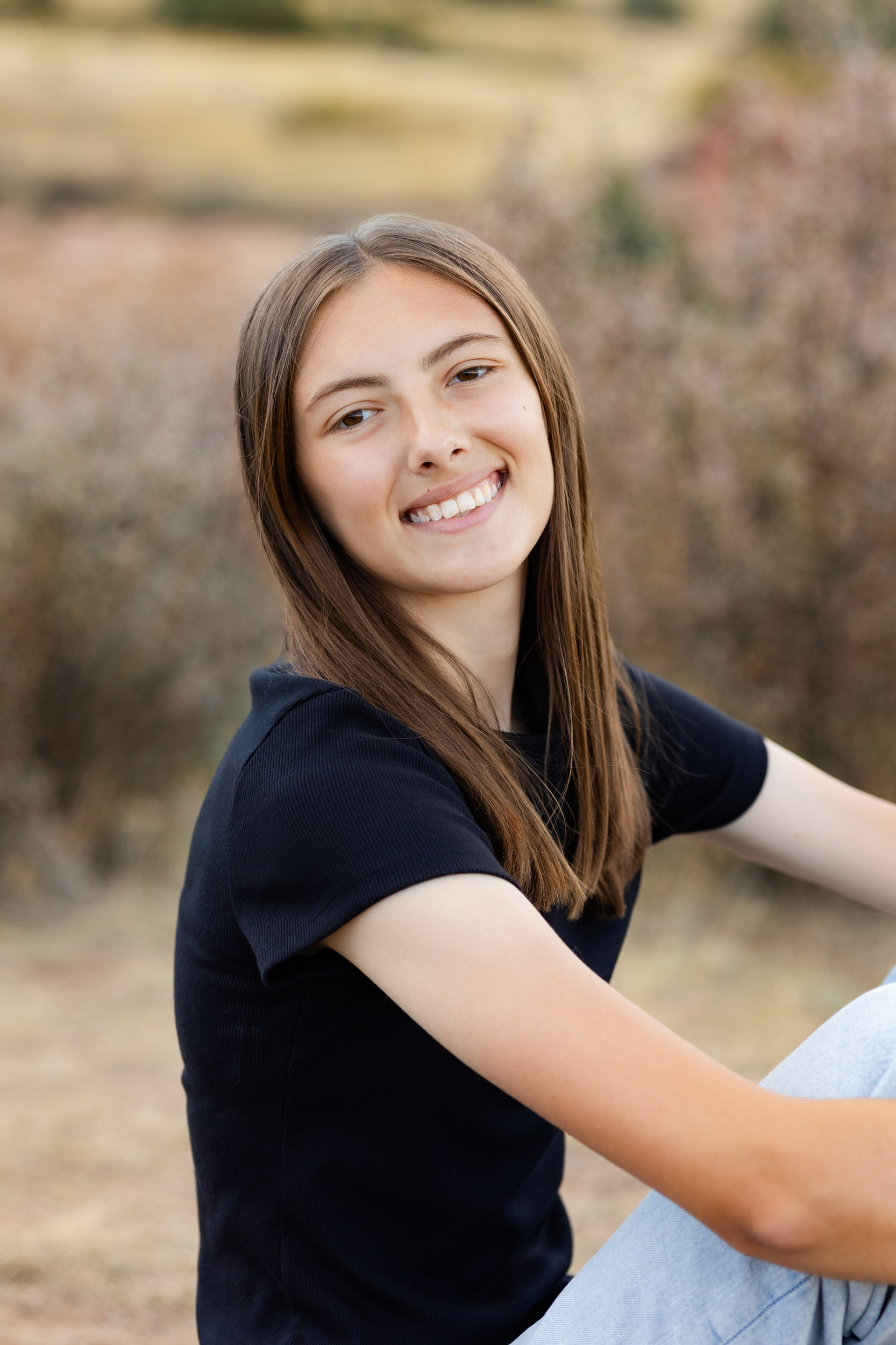 A young woman sits on a dirt path hugging her knees and smiles at the camera.