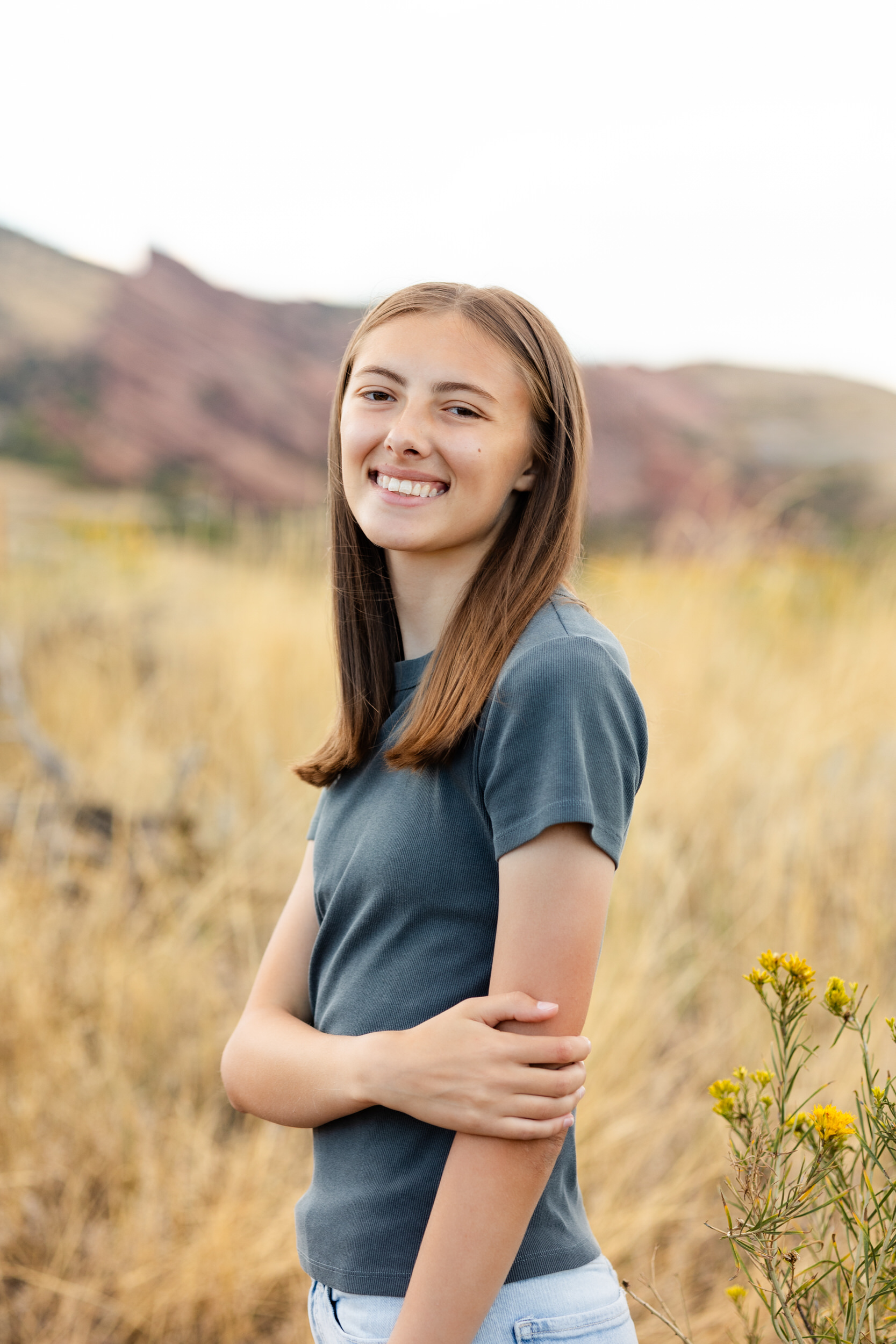 A young woman stands in a field of tall grass with yellow wildflowers and smiles at the camera.