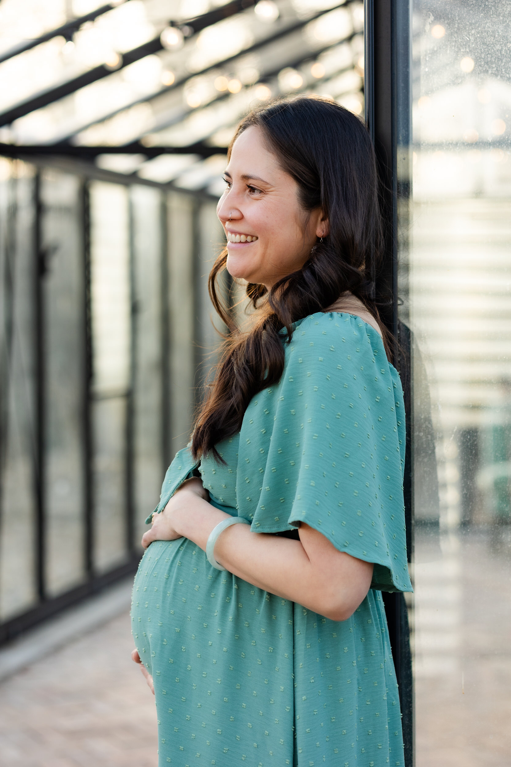 A pregnant woman leans on the doorway of a Denver greenhouse with both hands on her bump and smiles off-camera.