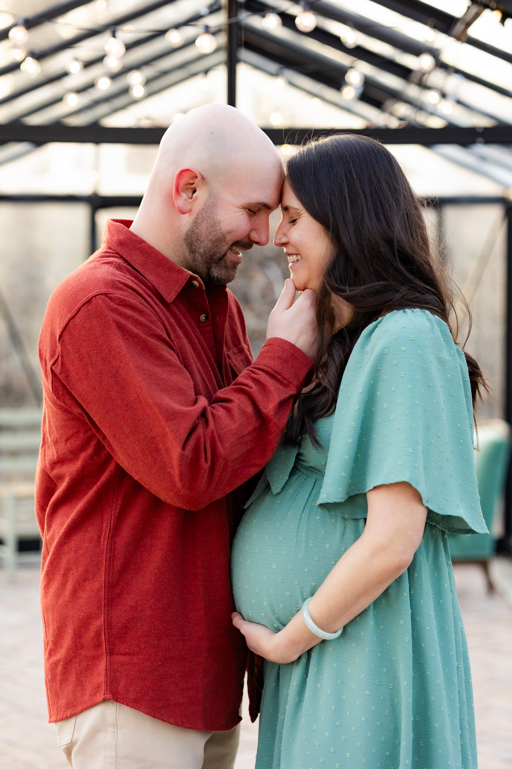 Husband and wife lean their foreheads together and smile as wife cradles her pregnant belly in her hands.