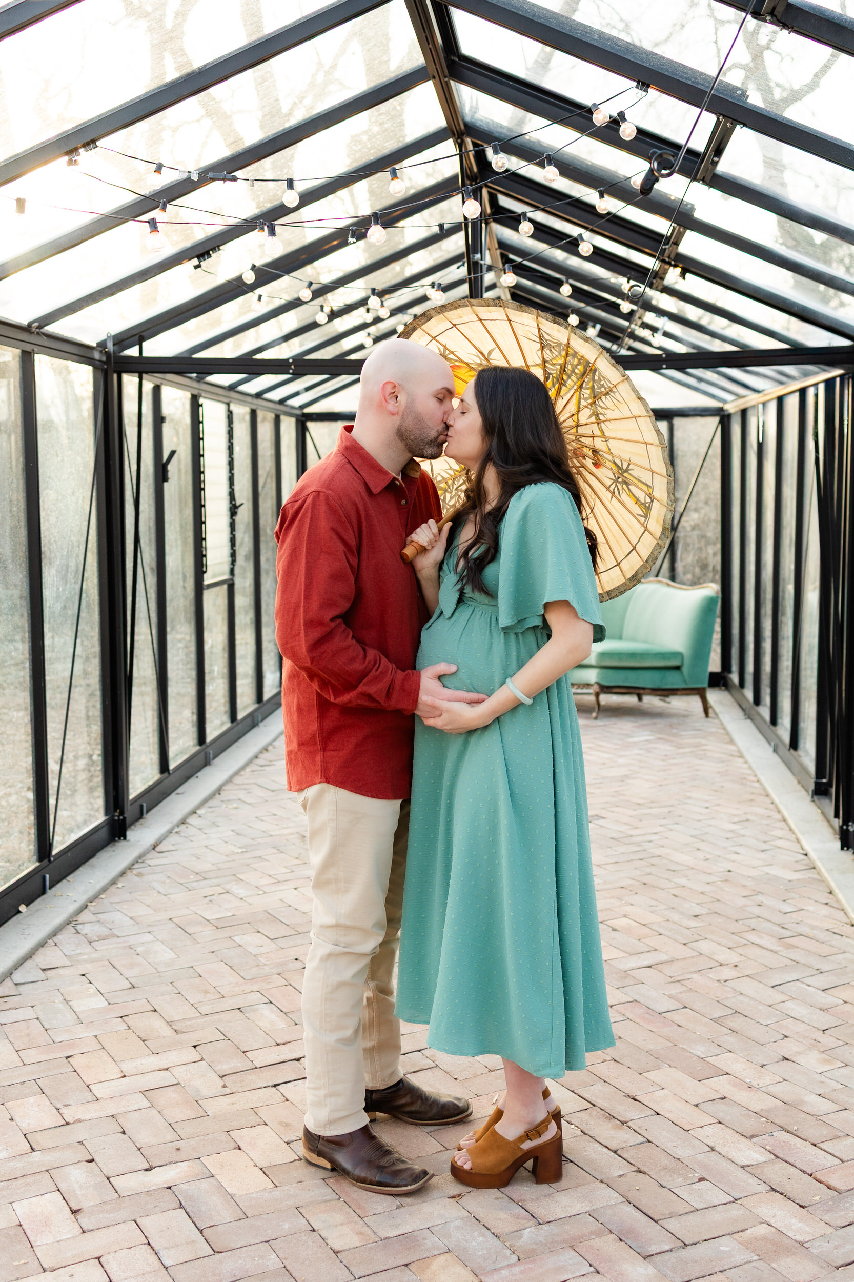 Husband and wife share a kiss beneath a parasol while standing in a greenhouse.