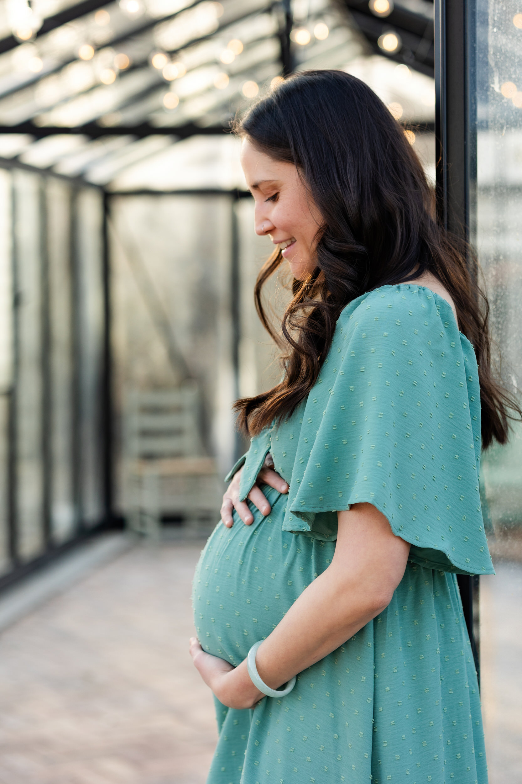 A pregnant woman leans on the doorway of a Denver greenhouse with both hands on her bump and smiles at her belly.