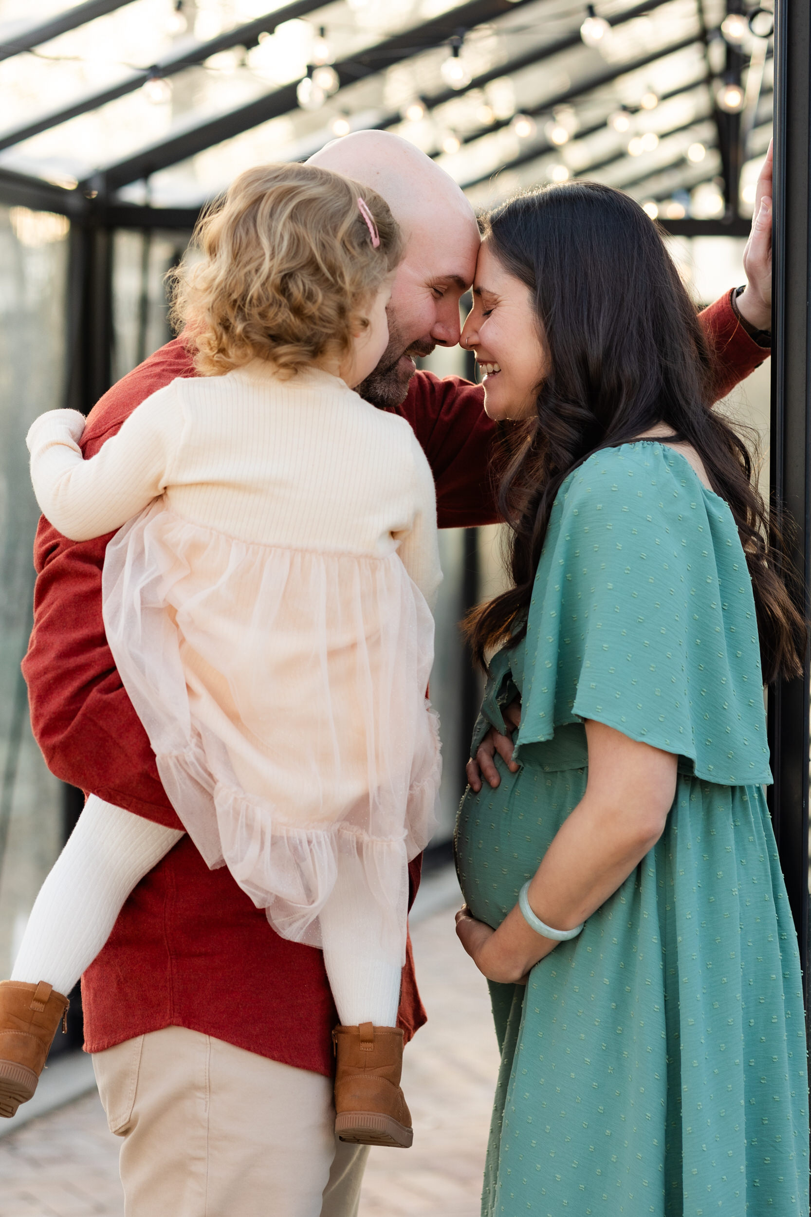 Husband and wife lean their foreheads together and smile while dad holds toddler daughter.
