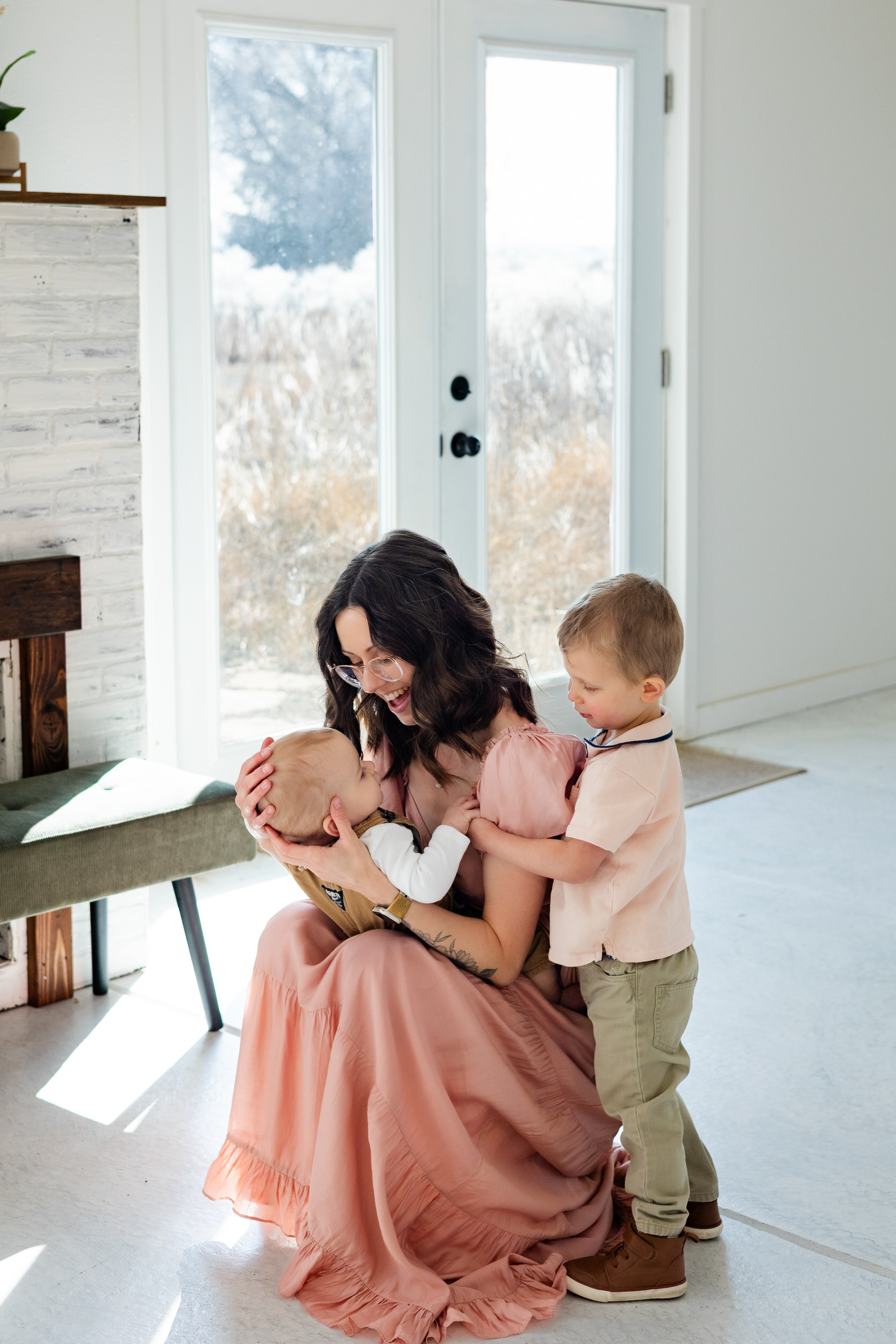 A mom holds her baby and smiles at him while older brother holds onto mom and also smiles at the baby.