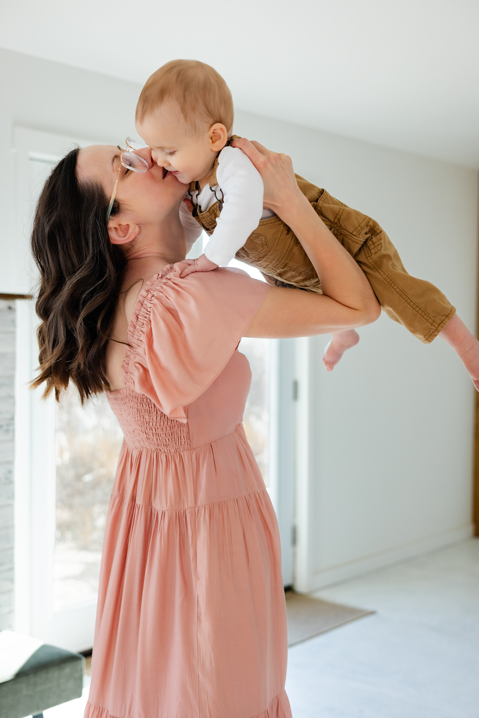 A mom holds her baby up in the air and kisses his cheek.