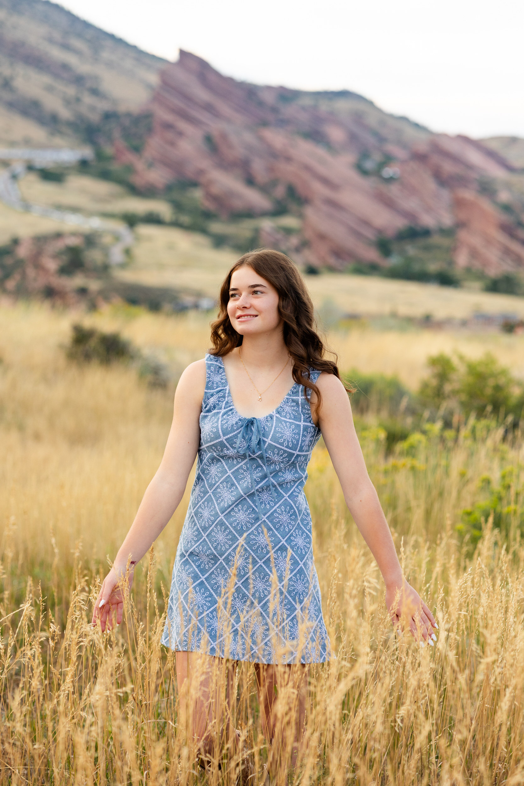 A young woman in a short blue dress walks through a field of tall grass and smiles off-camera.