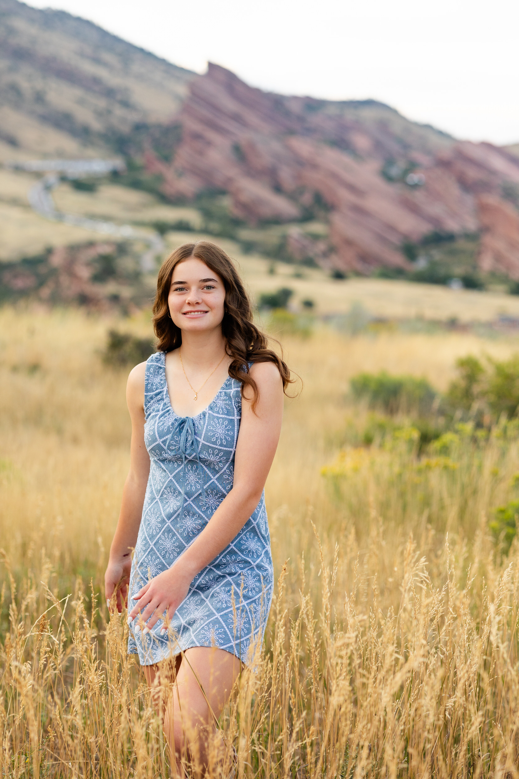 A young woman in a short blue dress walks through a field of tall grass and smiles at the camera.