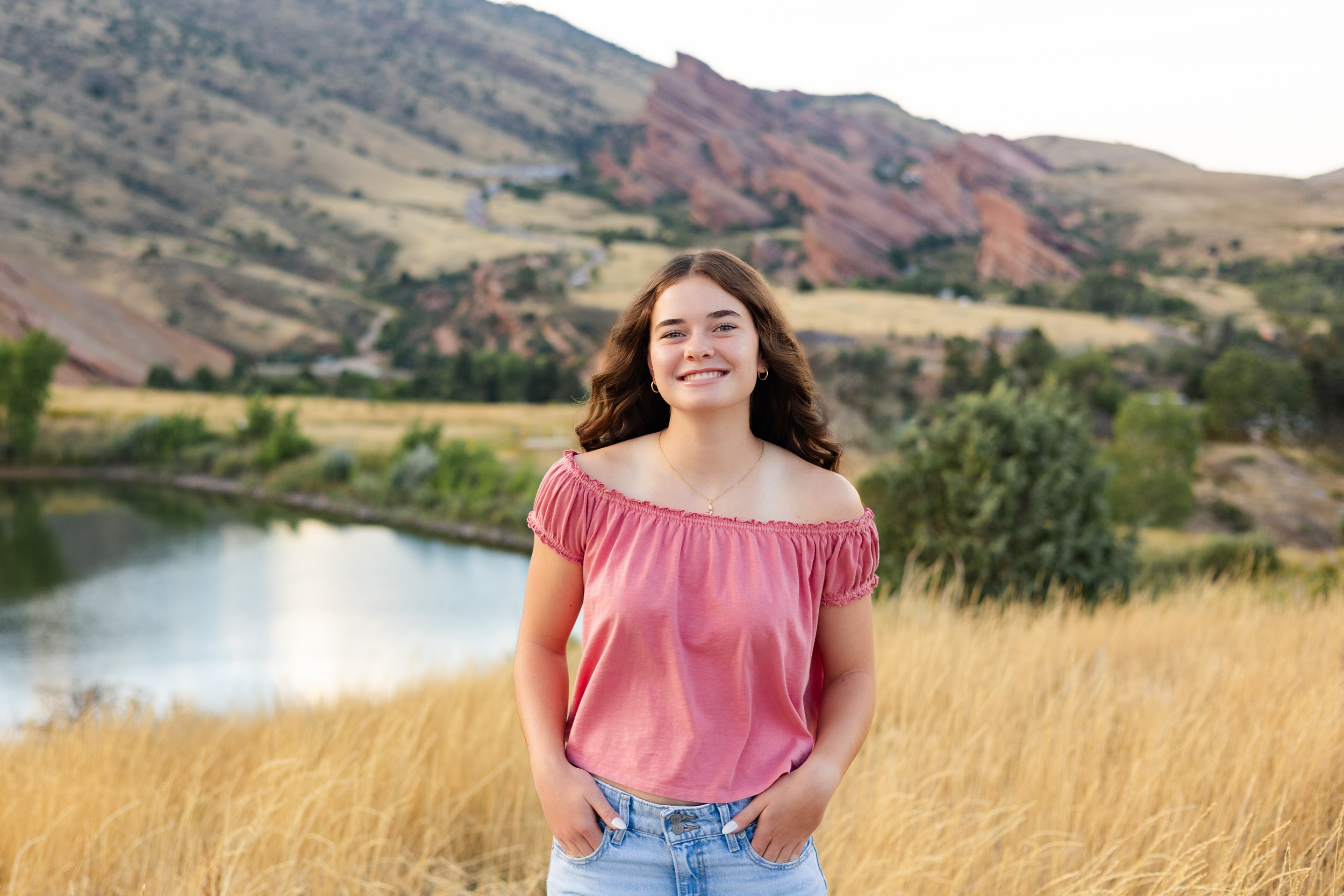 A young woman in a pink blouse stands in field with Red Rocks Amphitheater in the background and smiles at the camera.