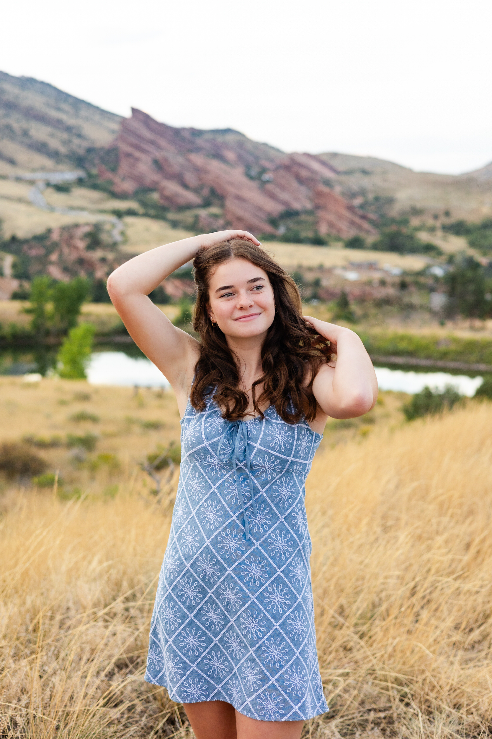 A young woman in a short blue dress runs both hands through her hair and smiles off-camera.