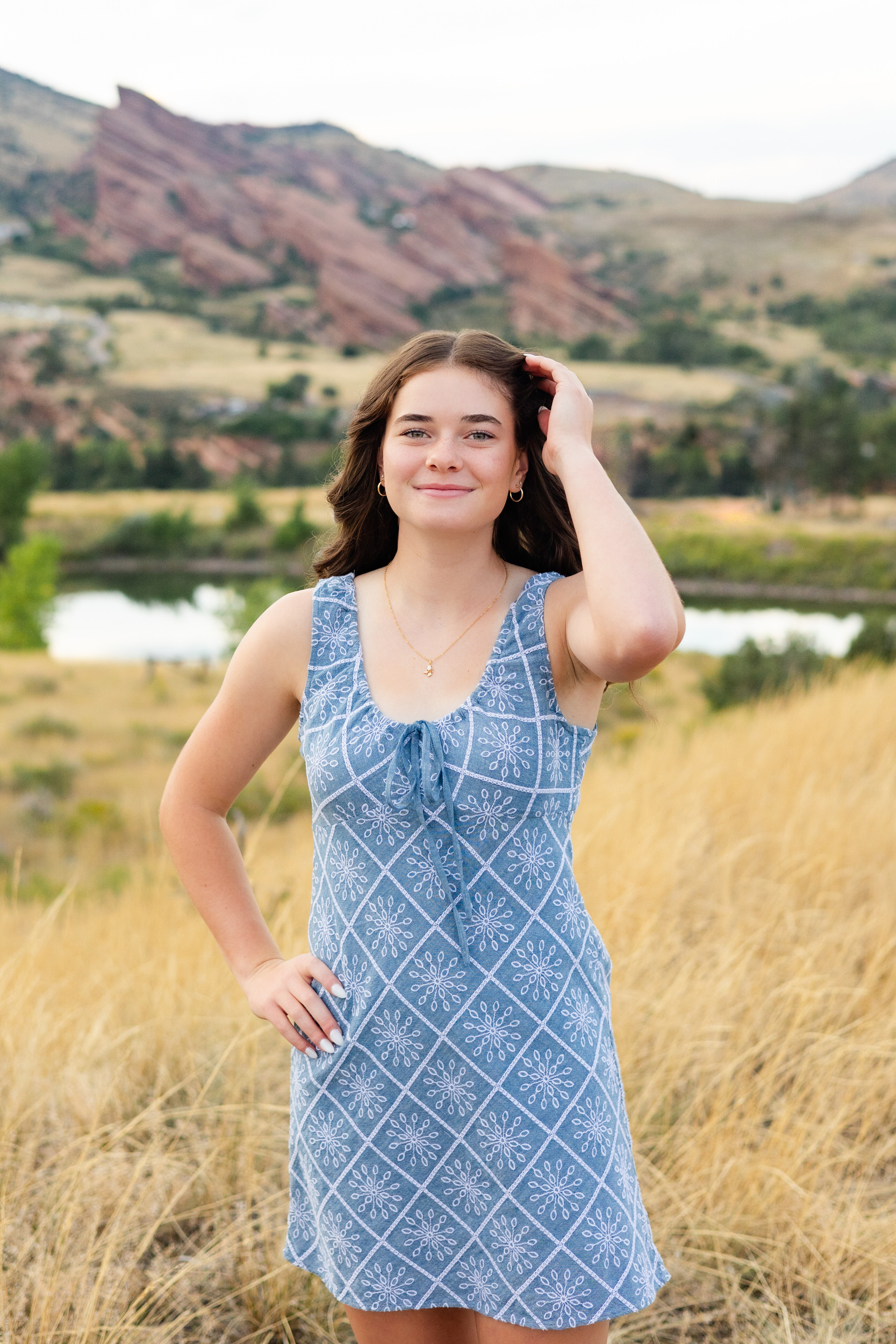 A young woman in a short blue dress runs one hand through her hair and smiles at the camera.