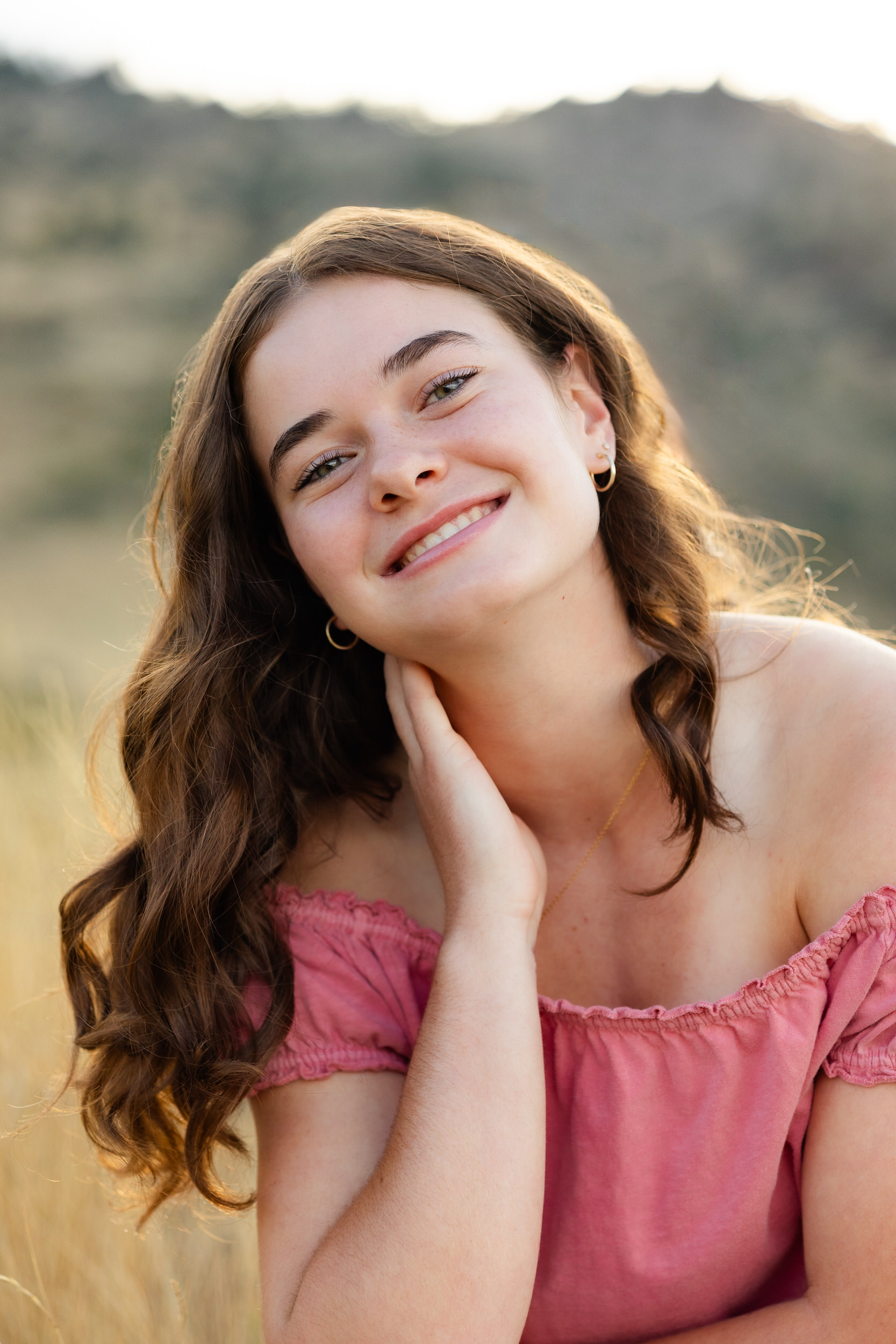 A young woman in a pink blouse squats in a field of tall grass resting her head on one hand and smiles at the camera.