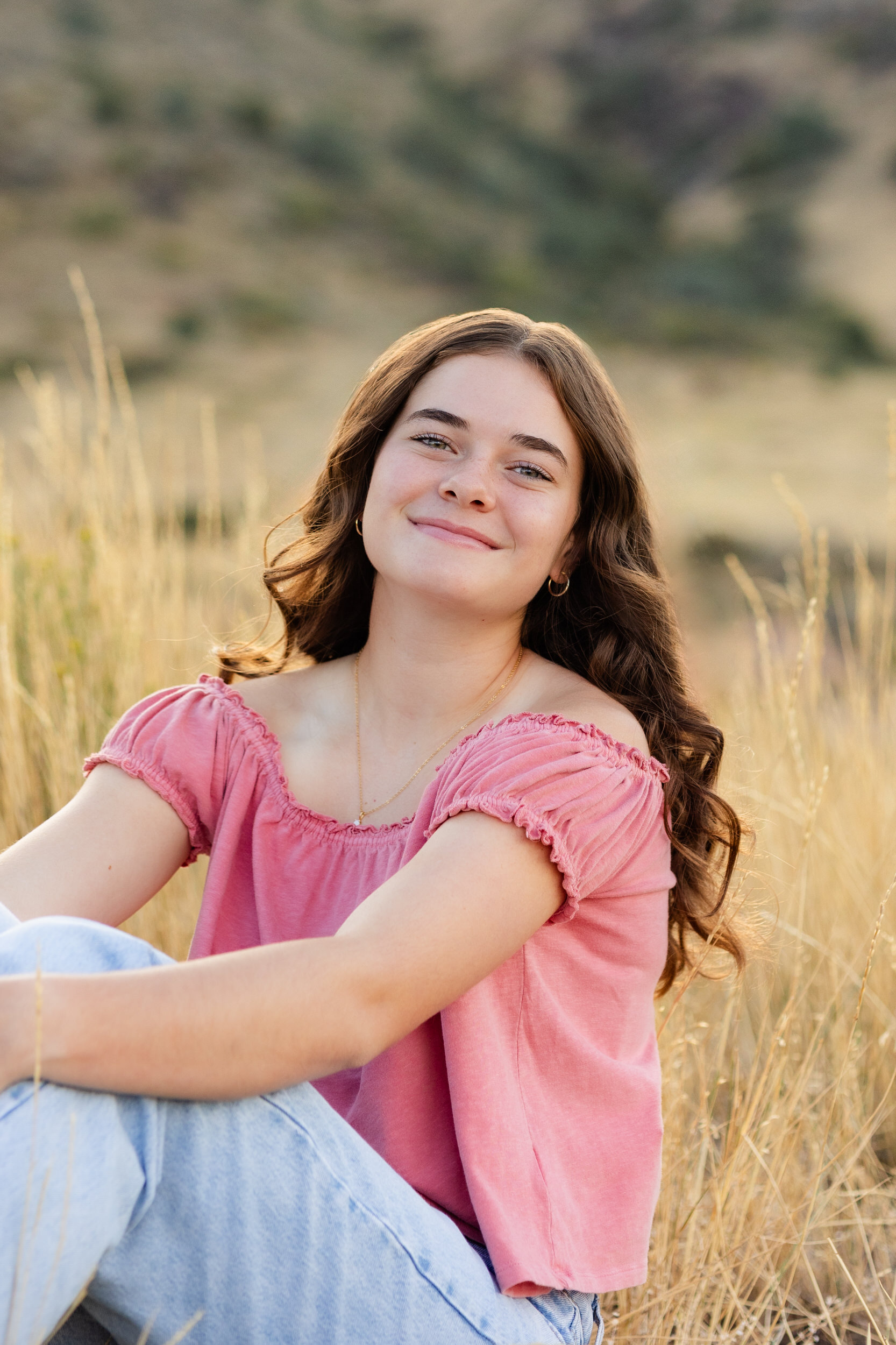 A young woman in a pink blouse and blue jeans sits in a field and smiles at the camera.