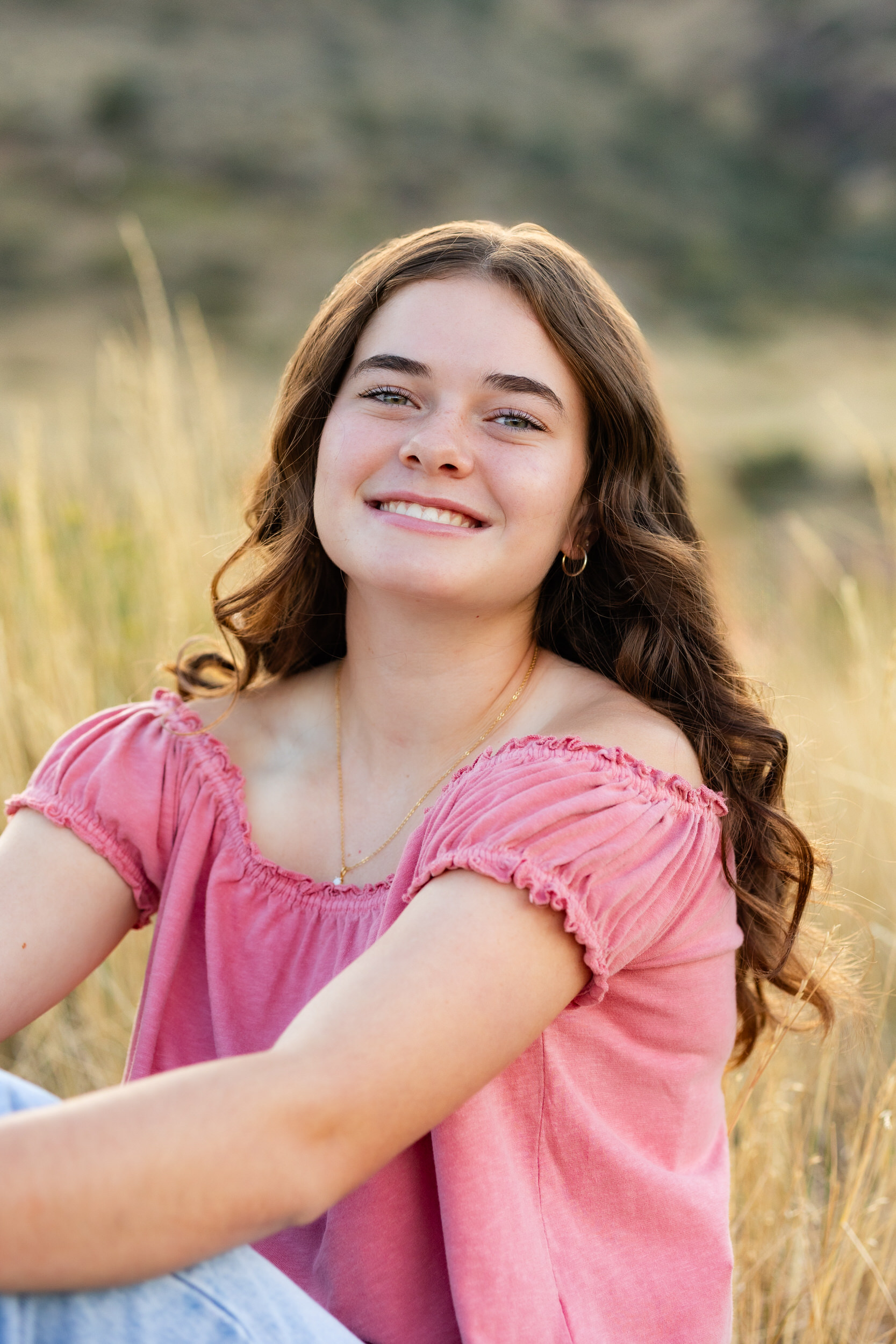 A young woman in a pink blouse and blue jeans sits in a field and smiles at the camera.