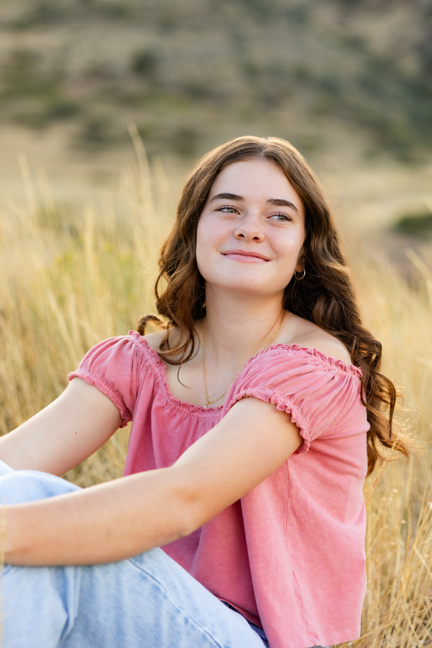 A young woman in a pink blouse and blue jeans sits in a field and smiles off-camera.