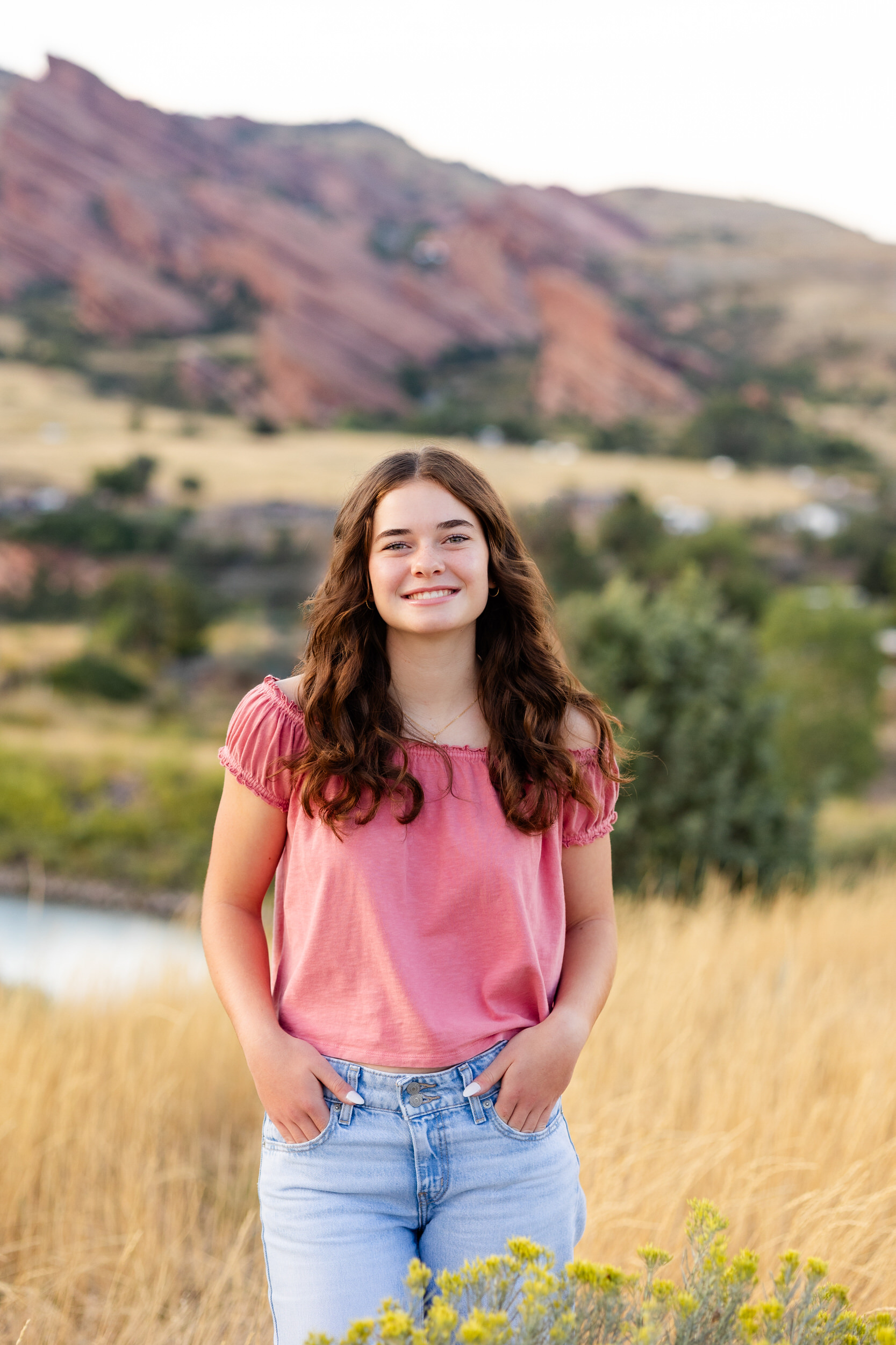 A young woman in a pink blouse and blue jeans stands in a field of tall grass and wildflowers and smiles at the camera.