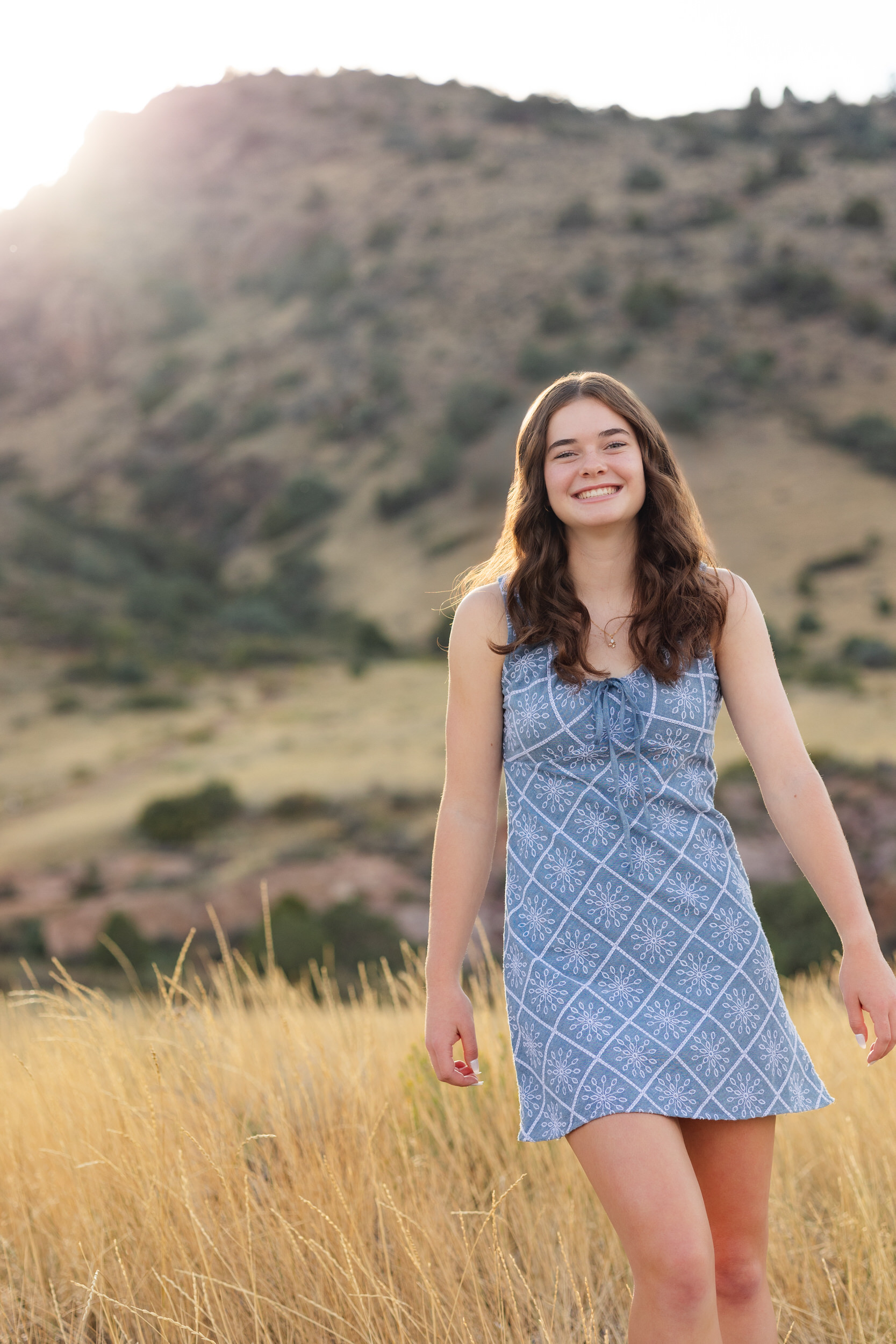 A young woman in a short blue dress walks toward the camera and smiles.