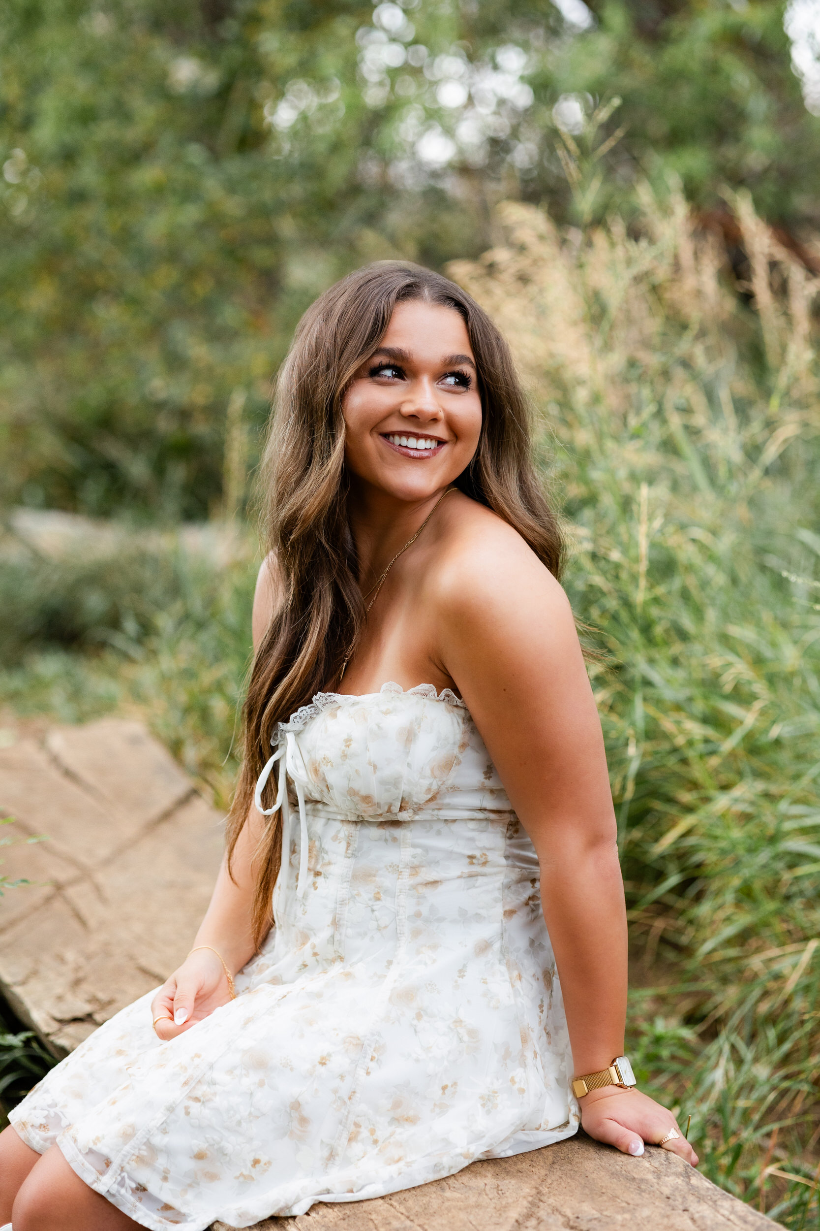 A young woman sits on a log bridge and smiles at the camera.