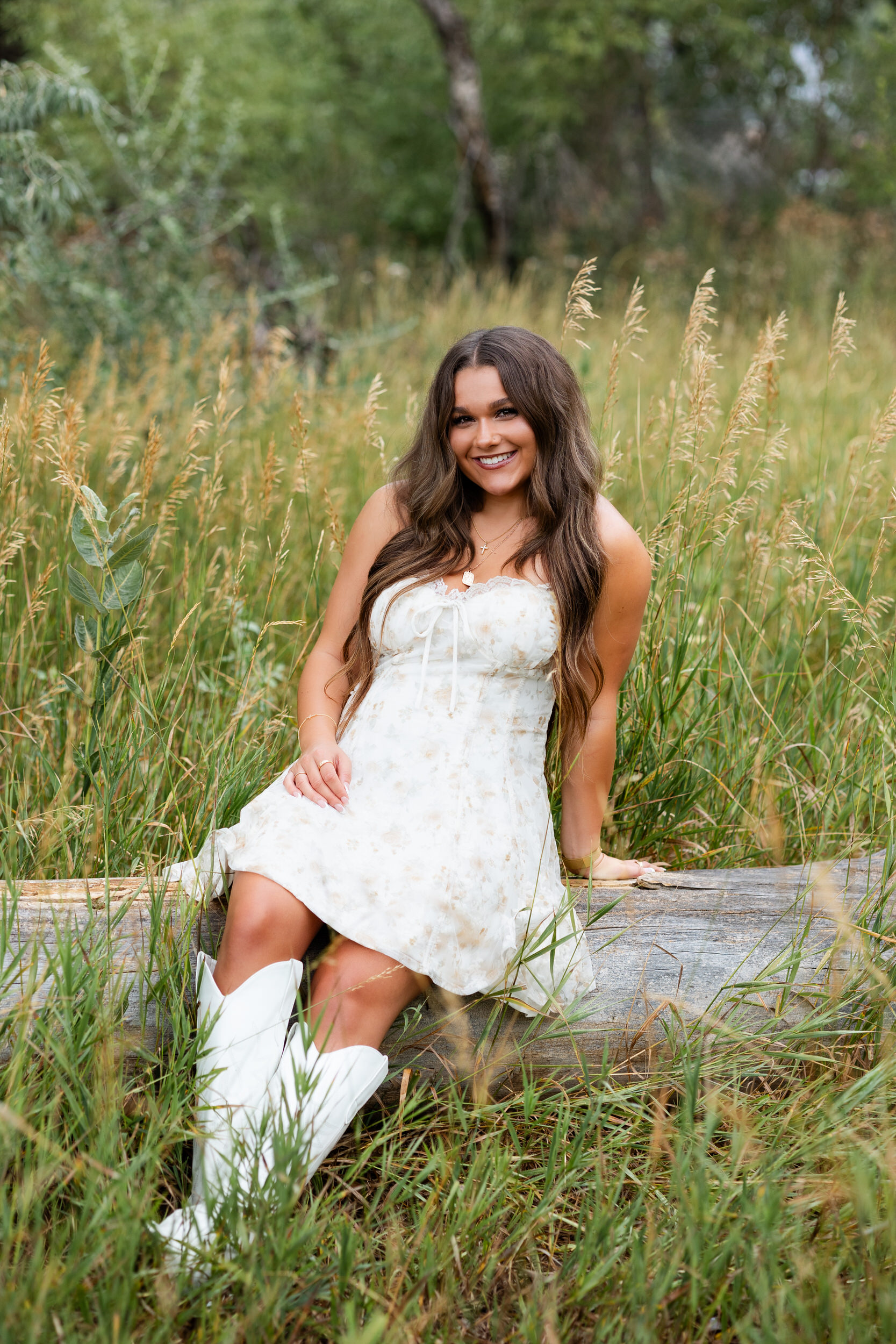 A young woman in a white dress sits on a log and smiles at the camera.