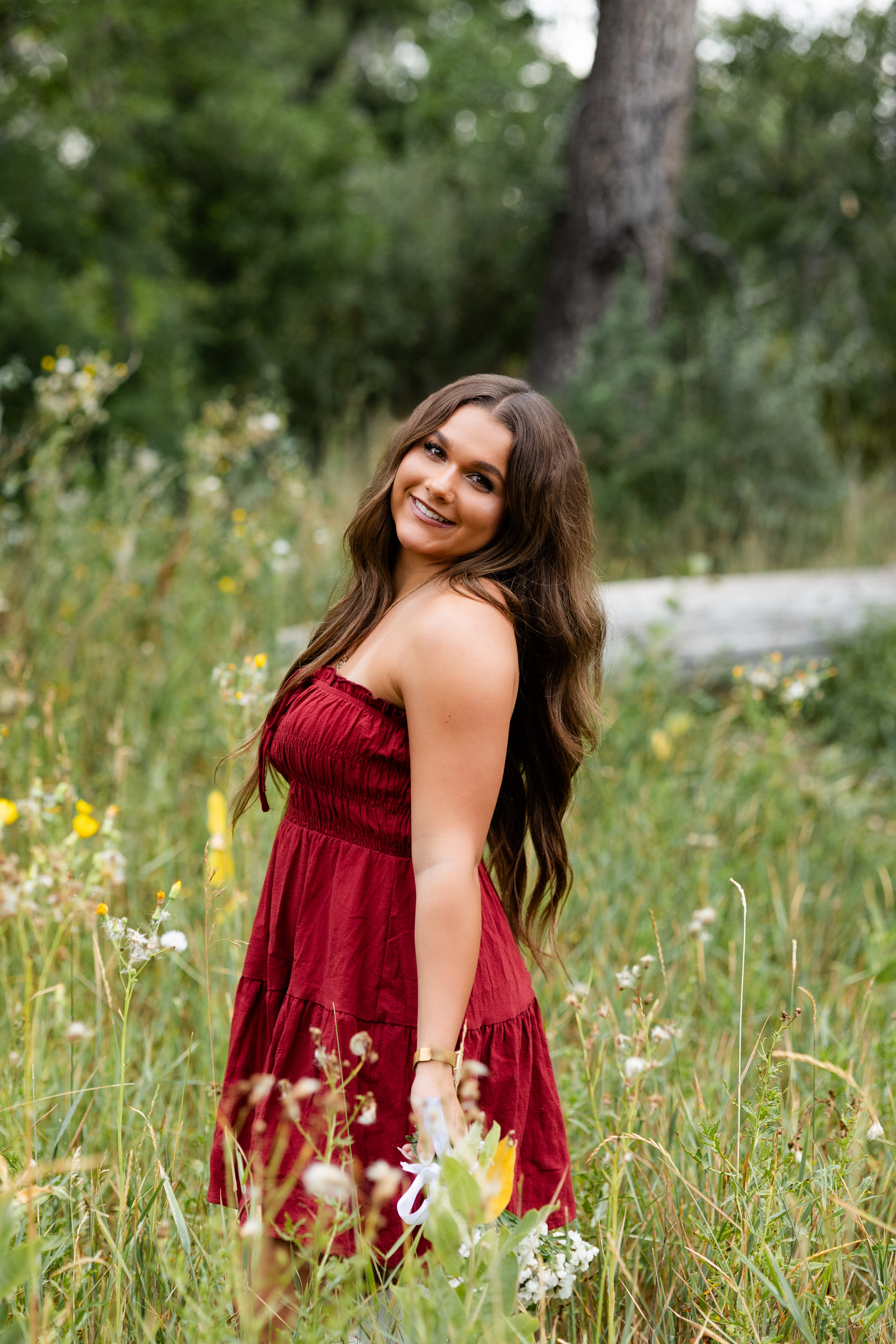 A young woman in a red dress stands in a field of tall grass and smiles at the camera.