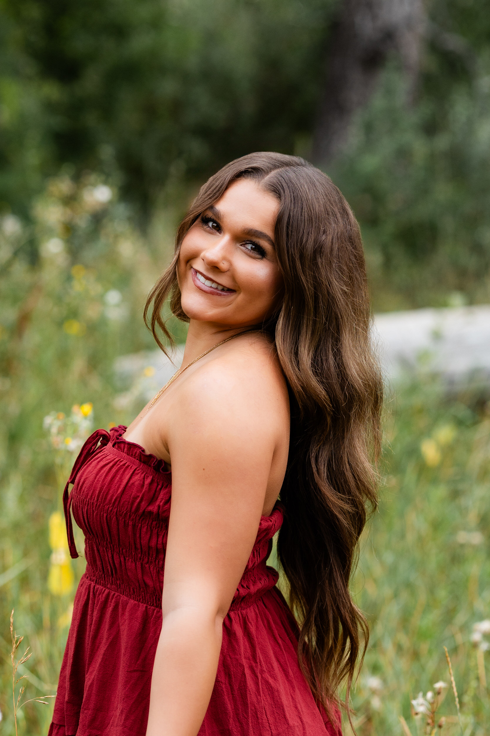 A young woman in a red dress stands in a field of tall grass and smiles at the camera.