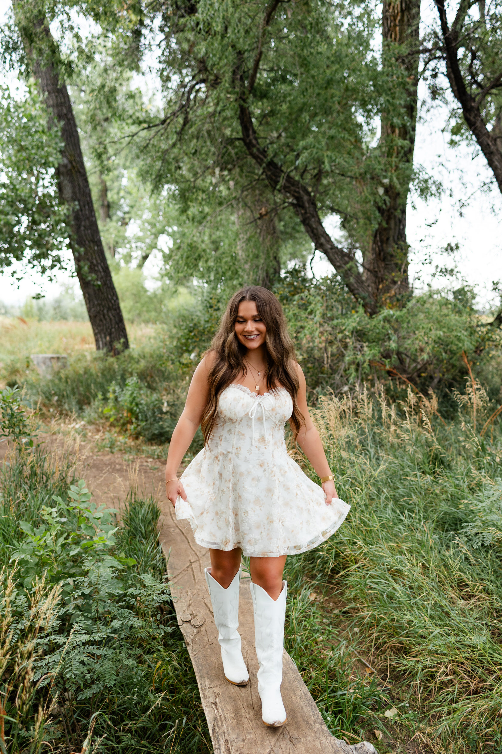 A young woman holds the hem of her dress and walks across a log bridge.
