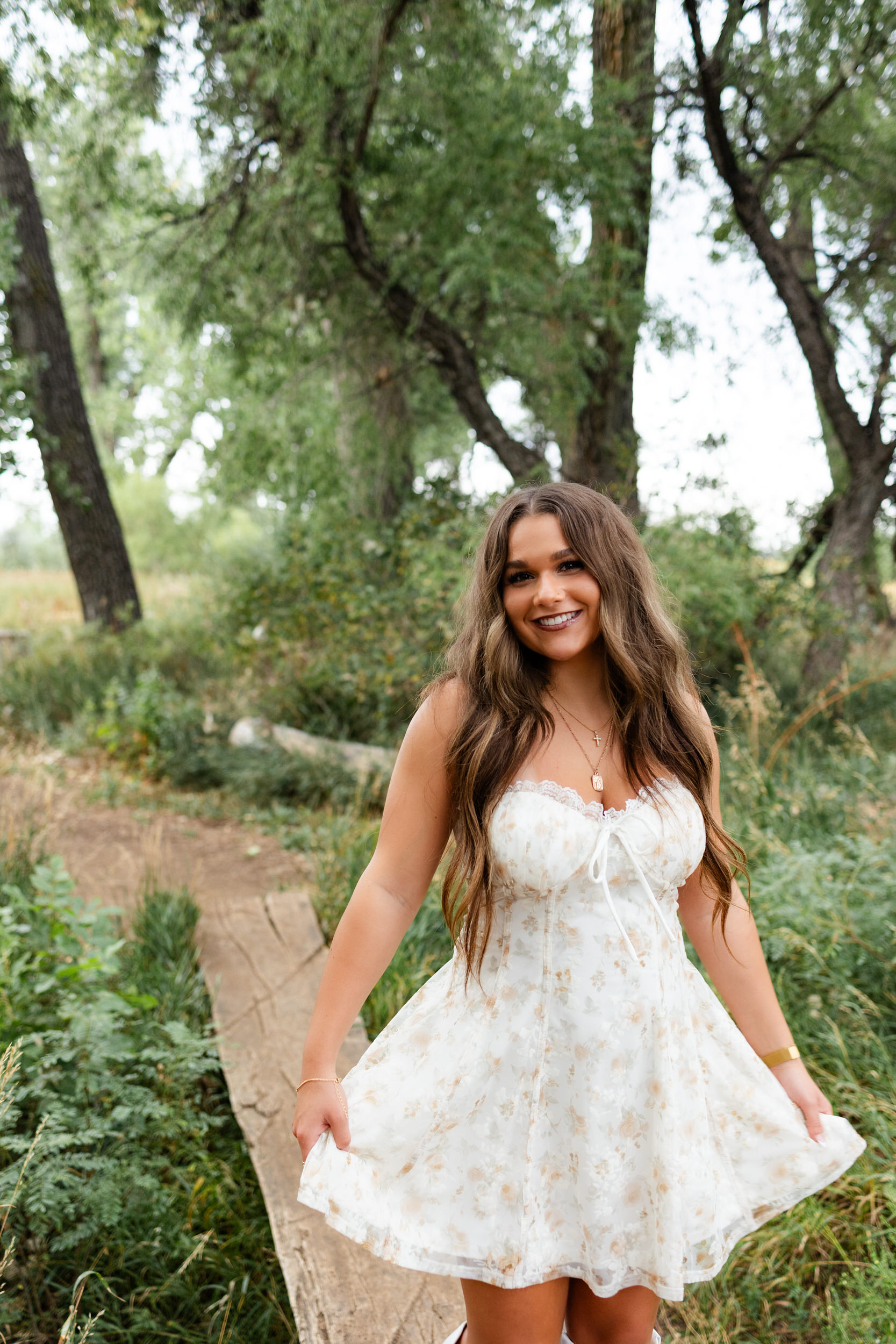 A young woman walks across a log bridge and holds onto the hem of her dress.