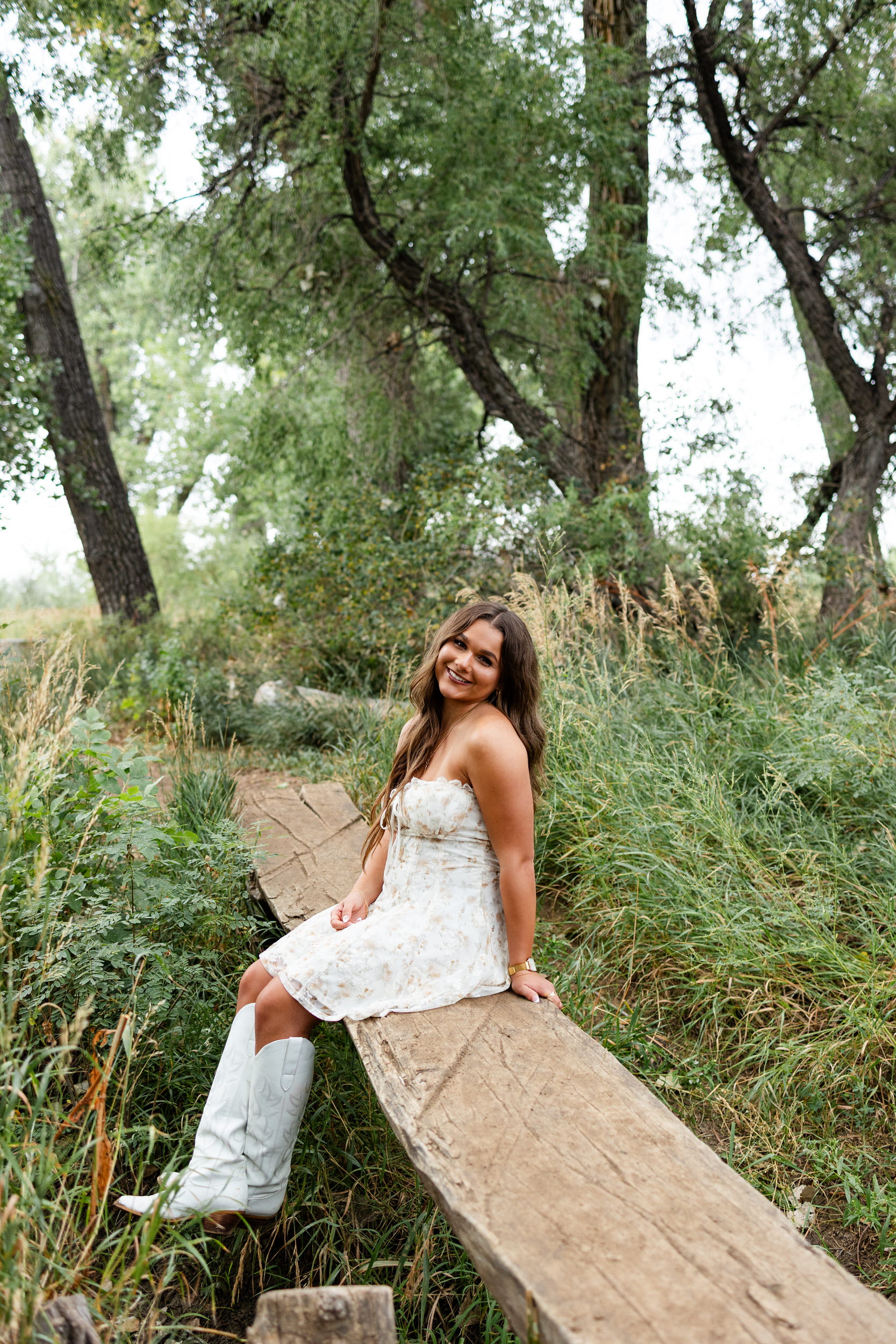 A young woman sits on a log bridge and smiles at the camera.