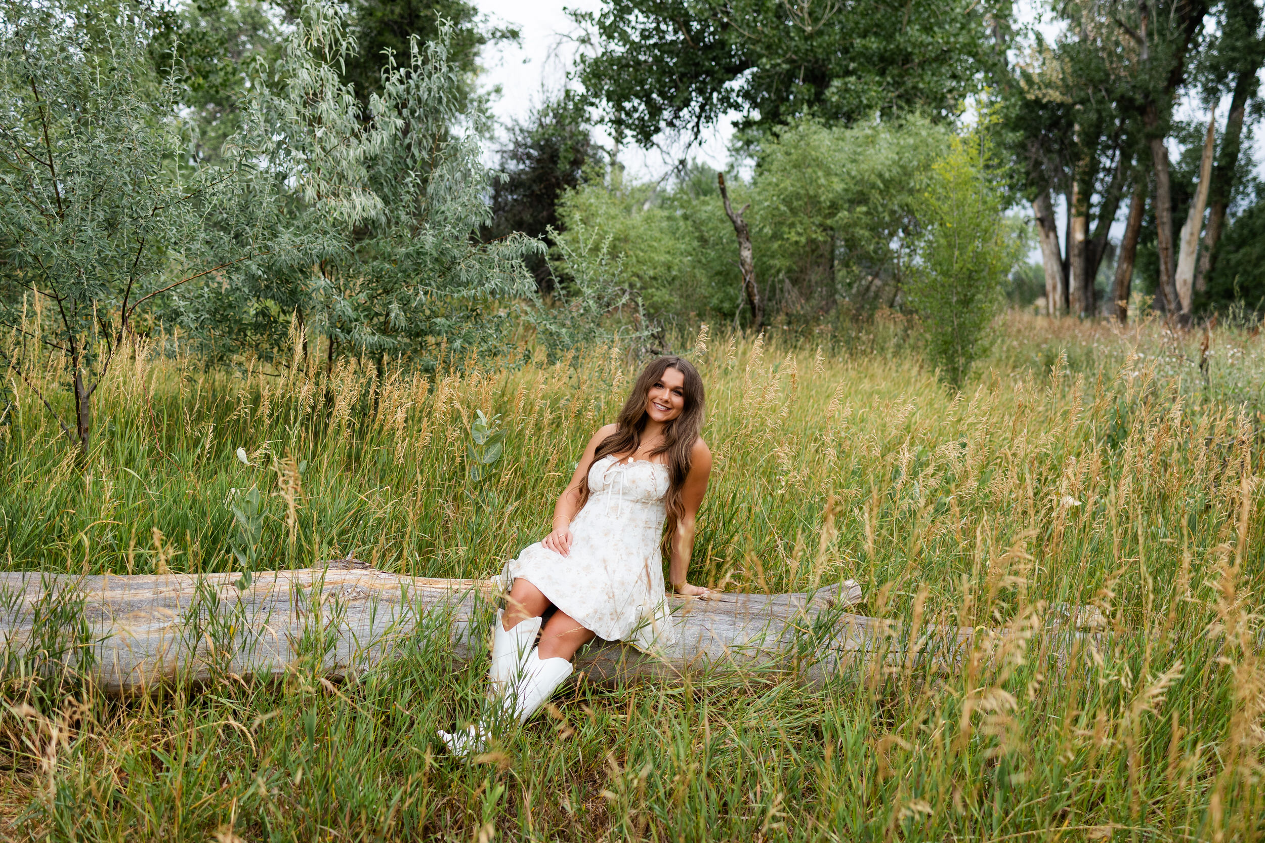 A young woman sits on a log in field of tall grass and smiles at the camera.