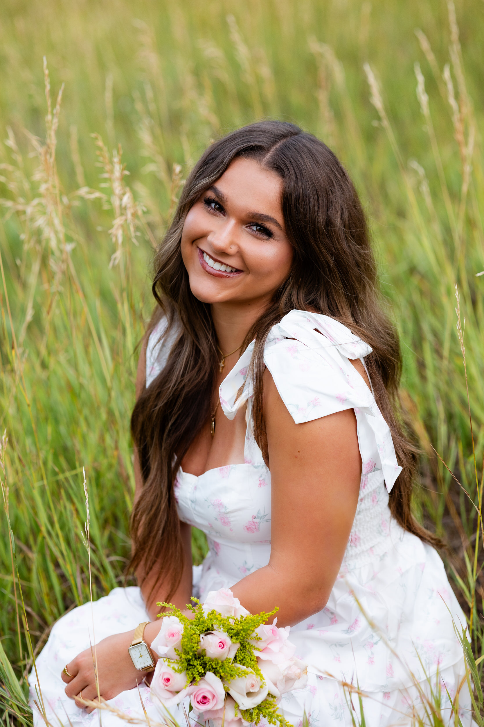 A young woman holding a bouquet of pink roses squats in a field of tall grass and smiles at the camera.