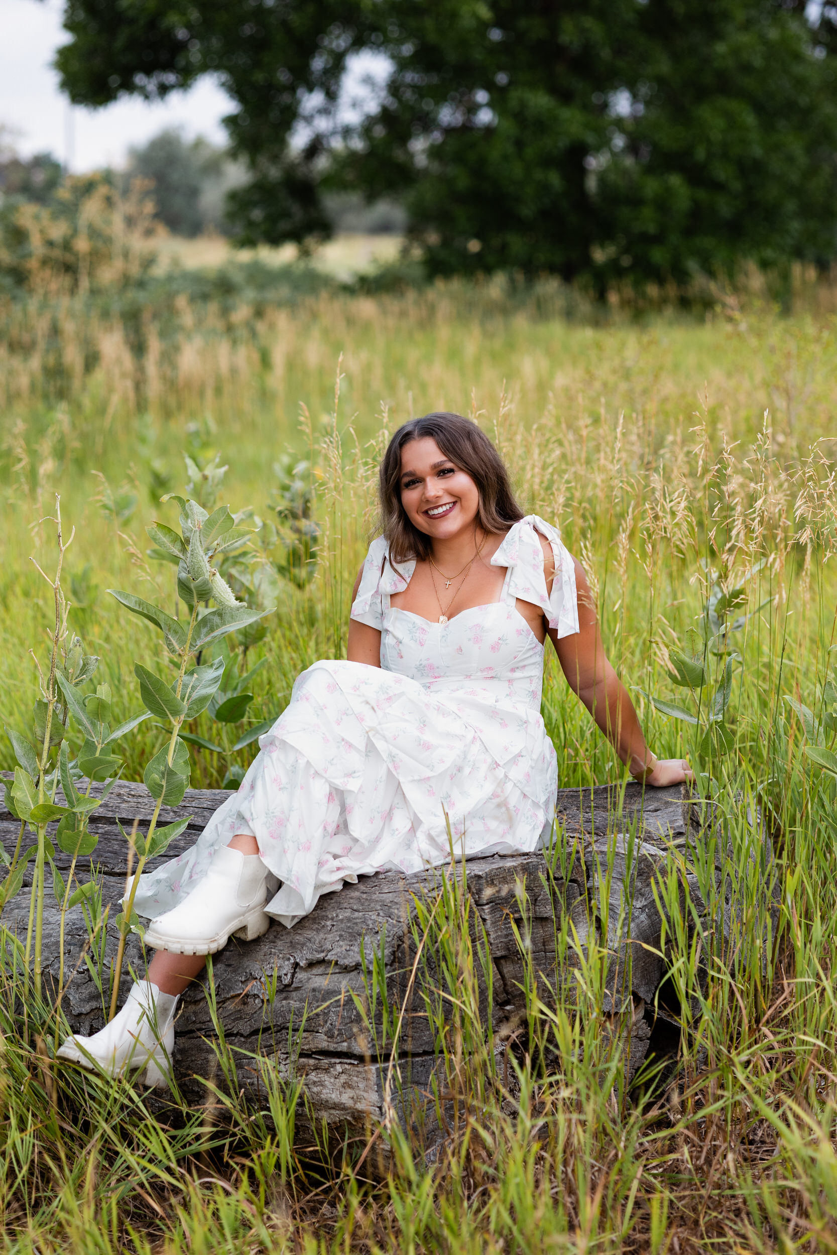 A young woman in a white dress sits on a log and smiles at the camera.
