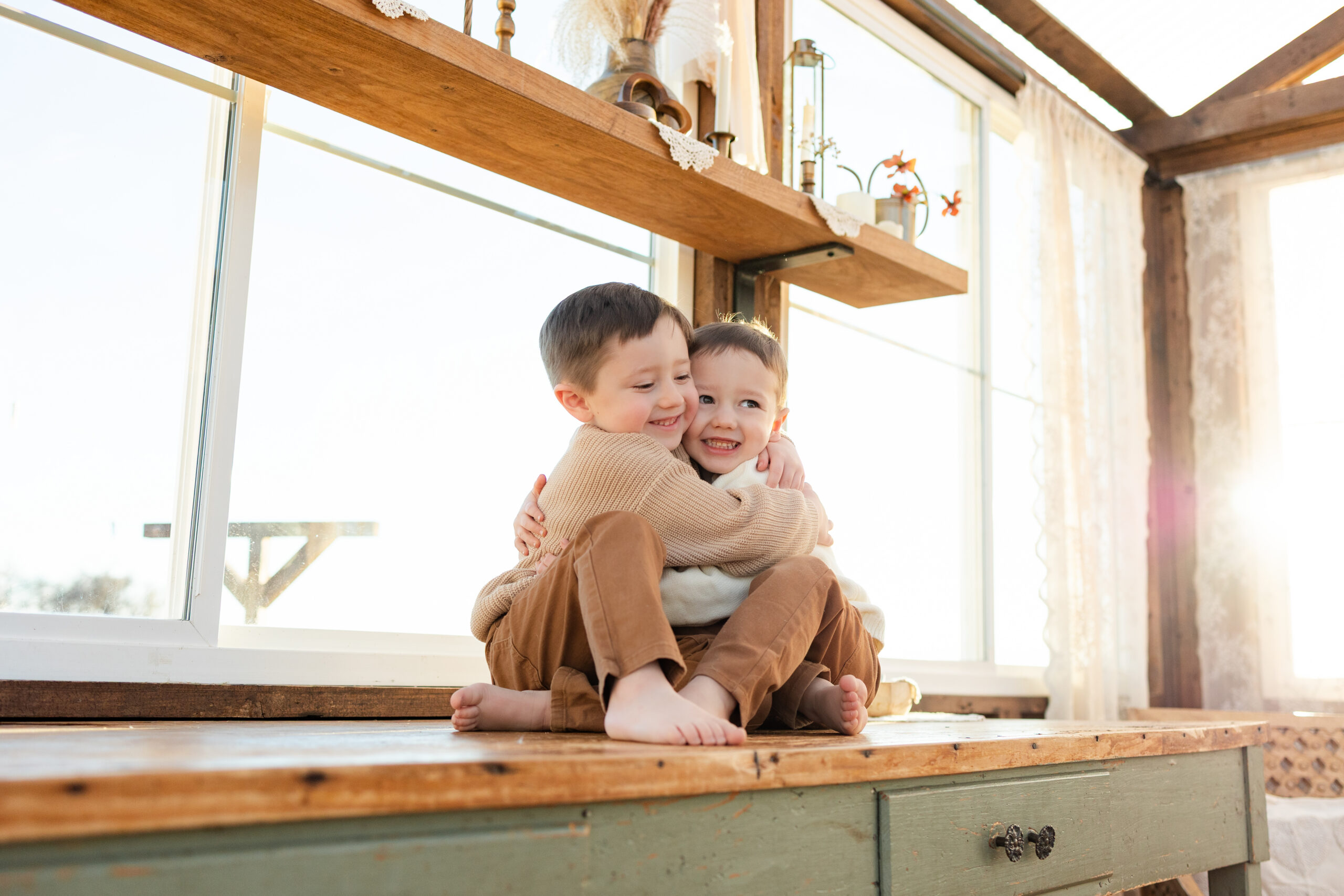 Two brothers sit on a table in a greenhouse hugging each other and smiling off-camera.