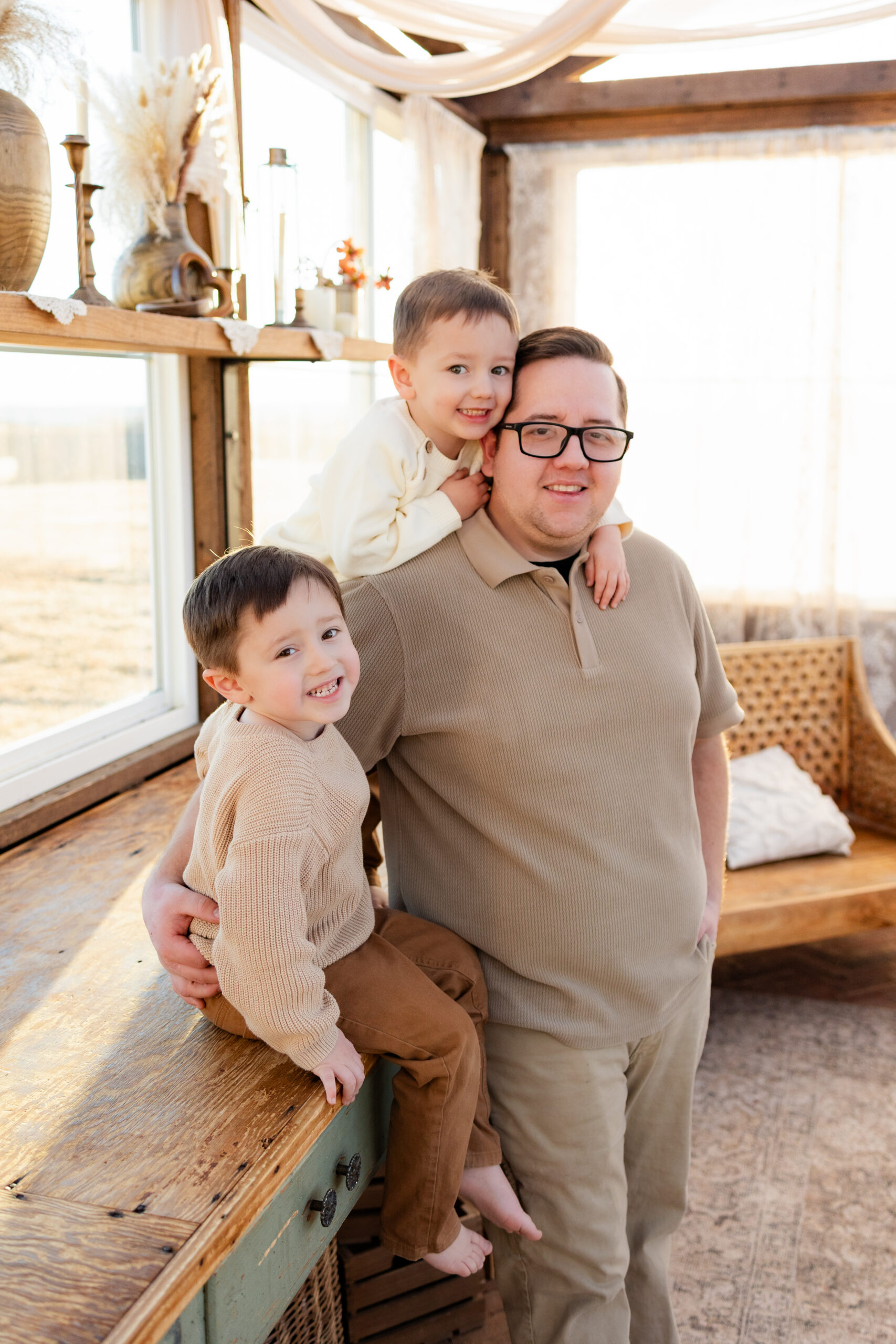 A dad stands next to a table in a greenhouse that his two young sons are sitting on and they all smile at the camera.