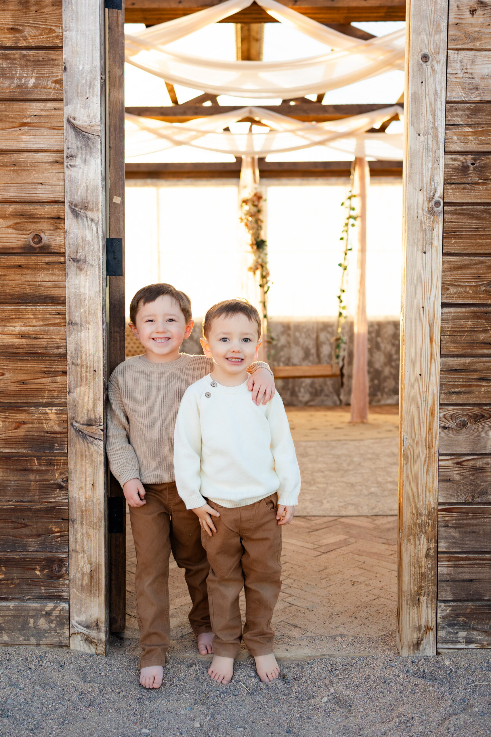 Two young brothers stand in the doorway of a greenhouse with their arms around each other's shoulders smiling at the camera.