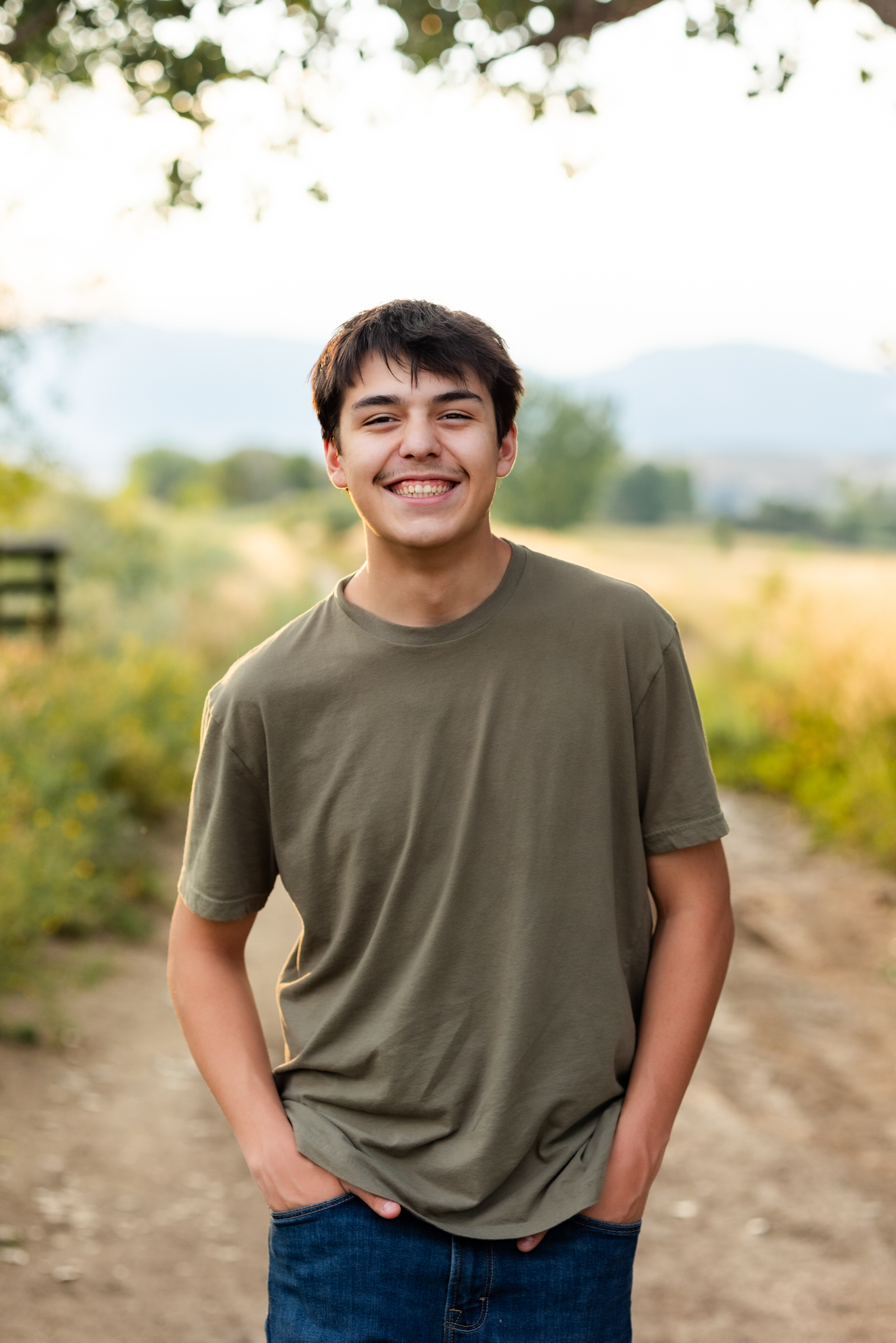 A young man walks on a dirt trail with his hands in his pockets smiling at the camera.