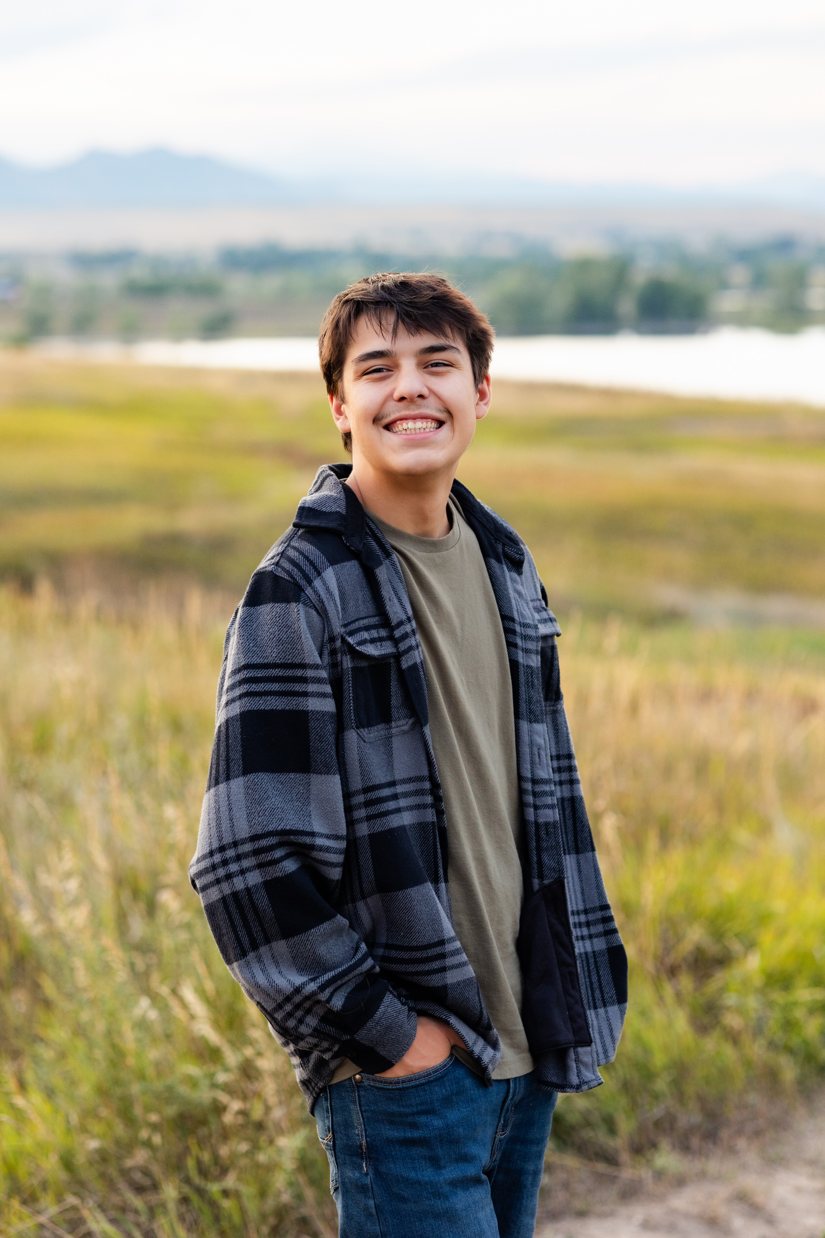 A young man in a plaid shirt stands on a dirt trail in front of a lake with his hands in his pockets smiling at the camera.