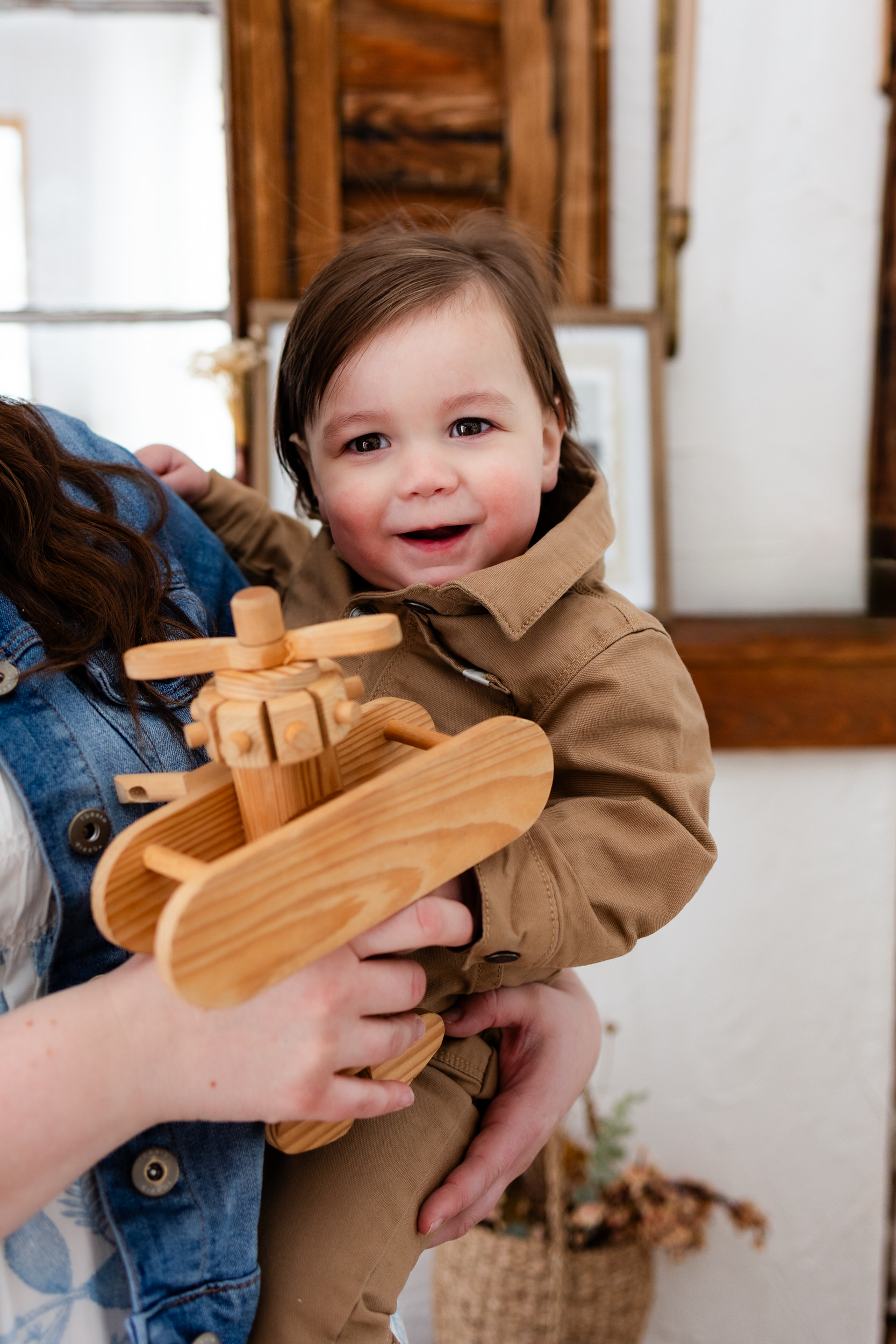 Mom rests toddler boy on her hip as he smiles at the camera and she holds a toy airplane for him.