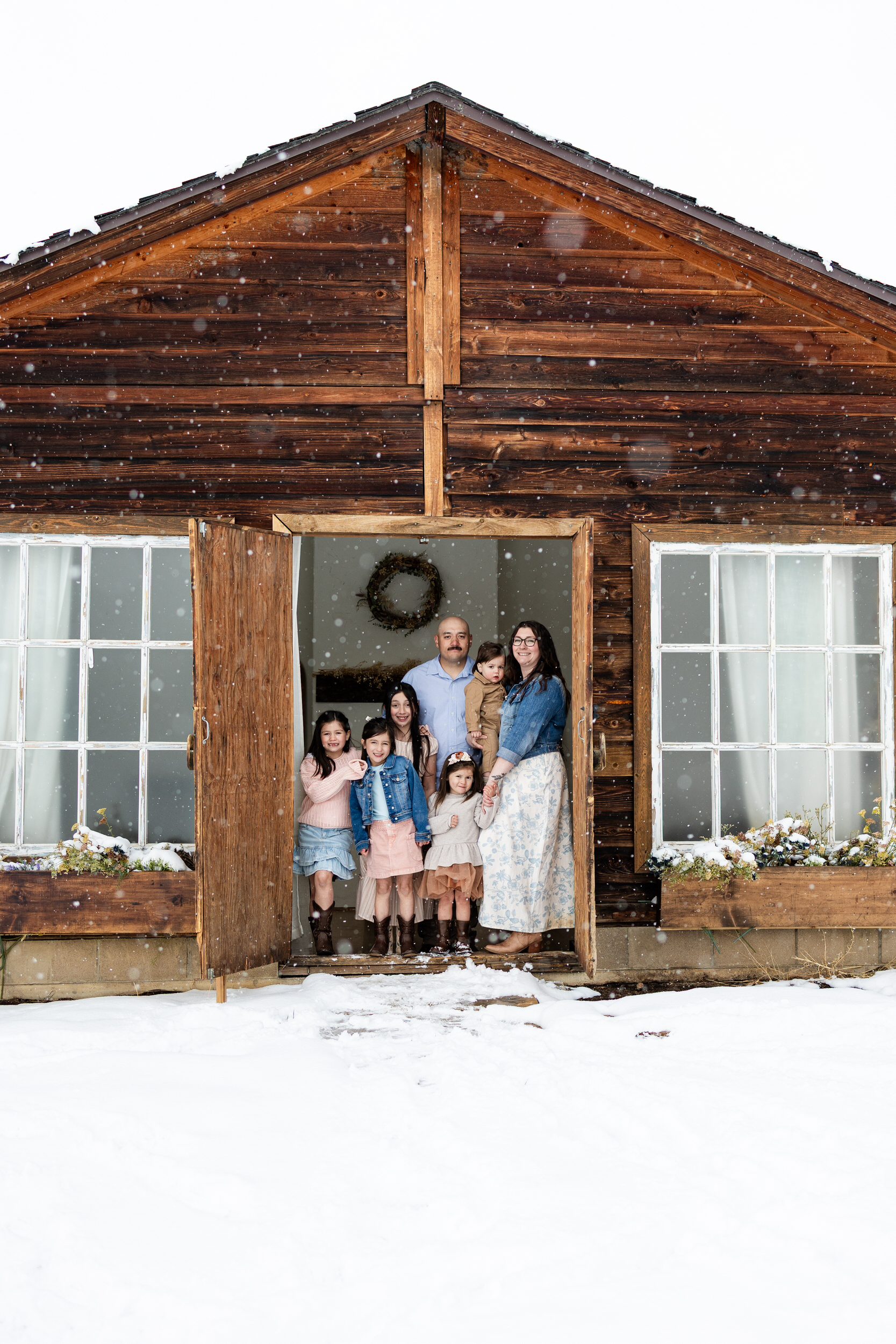 Mom and dad stand with their five young kids in the doorway of a cottage as it snows outside.