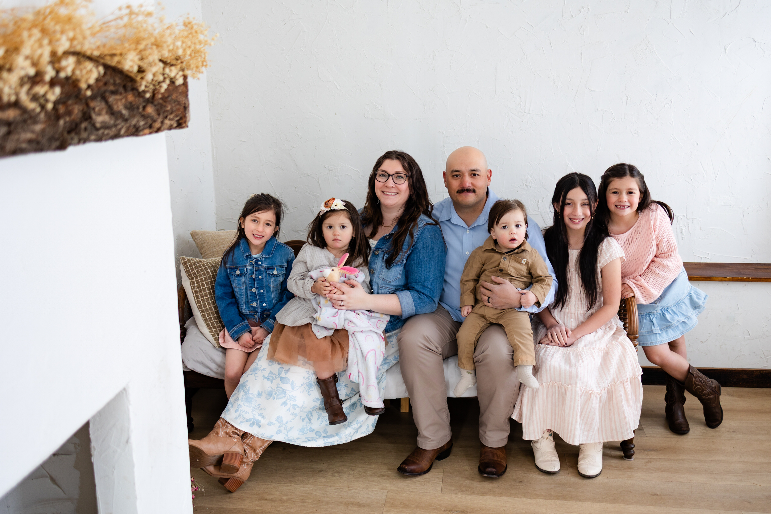 A mom and dad sit with their five young kids on a bench in a cottage and smile at the camera.