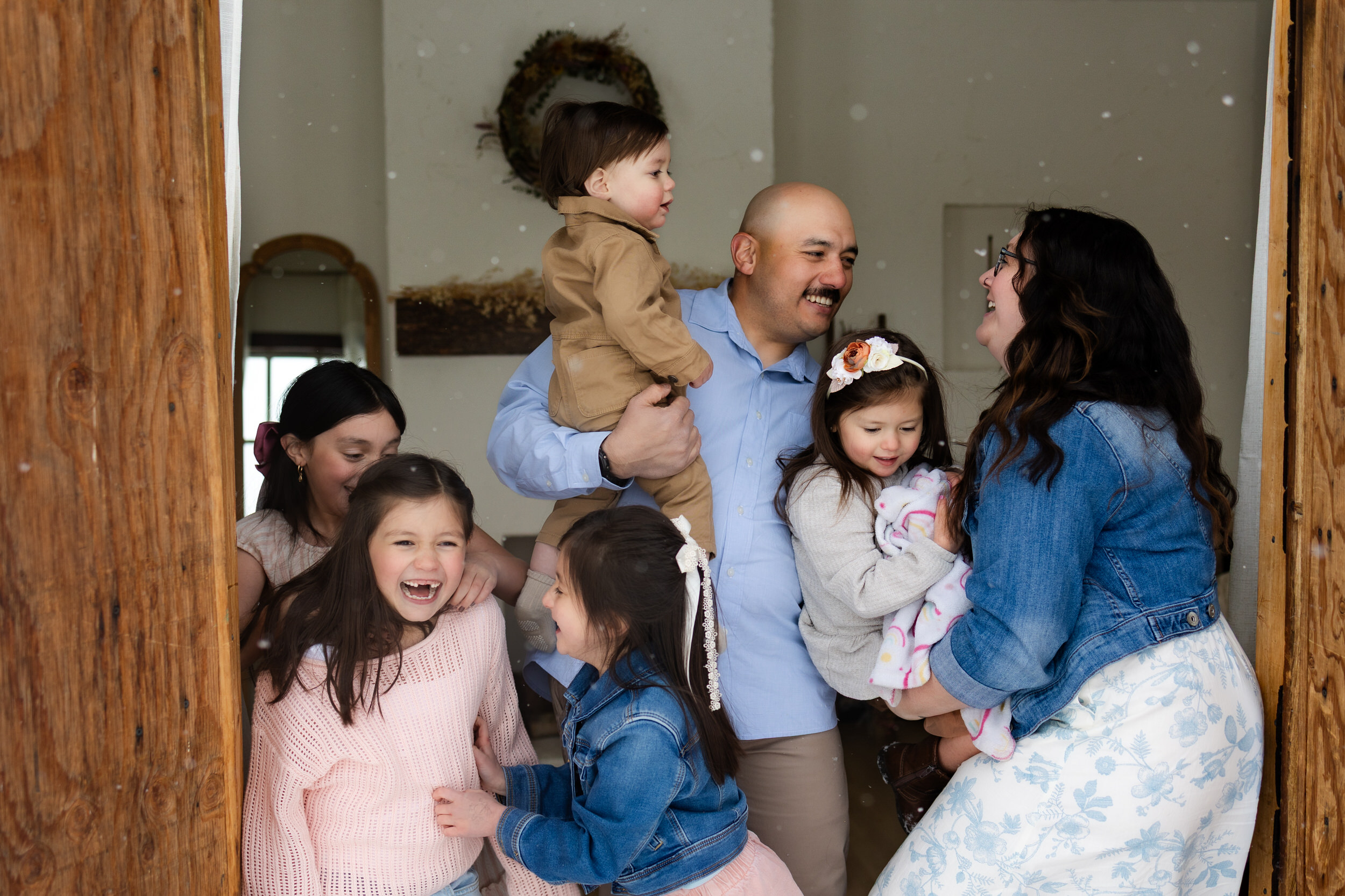 Mom and dad play with their five young kids in the doorway of a cottage as it snows outside.