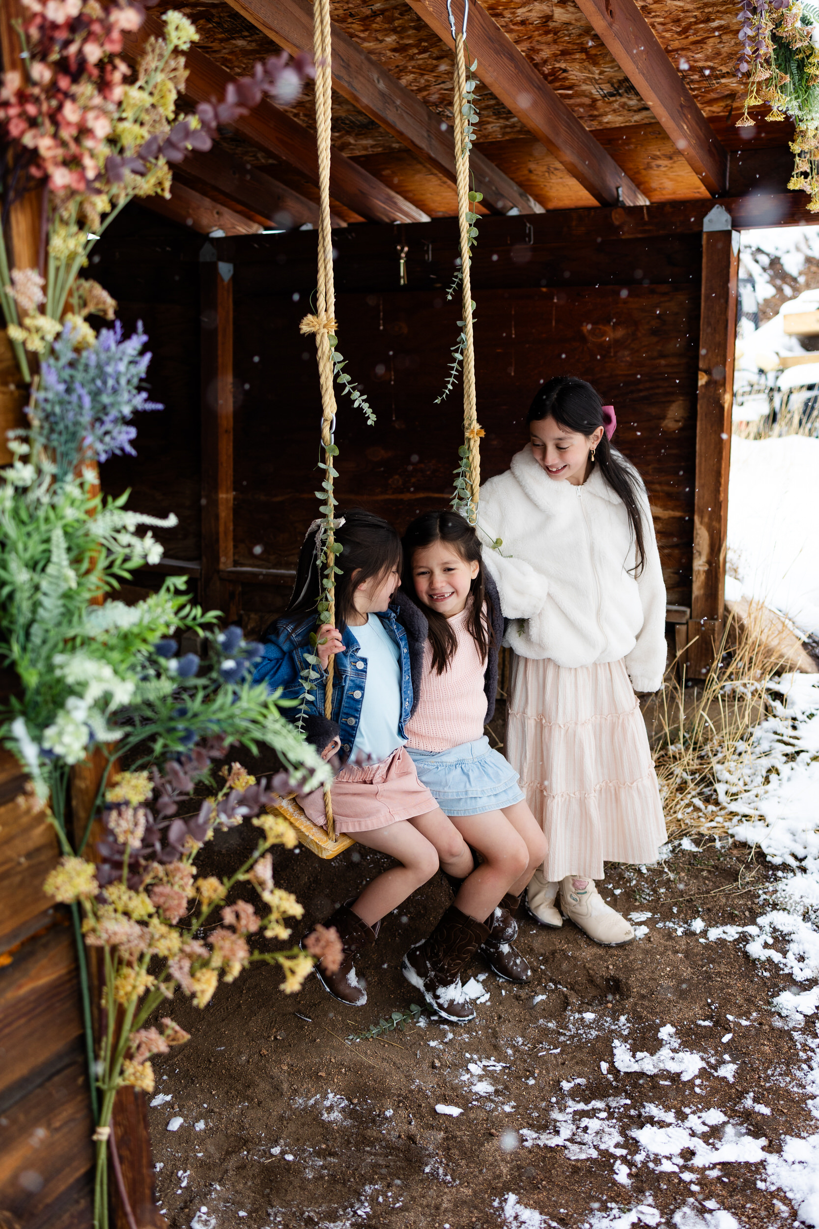 Two sisters sit on a swing while the oldest sister stands beside them holding one of the ropes of the swing.