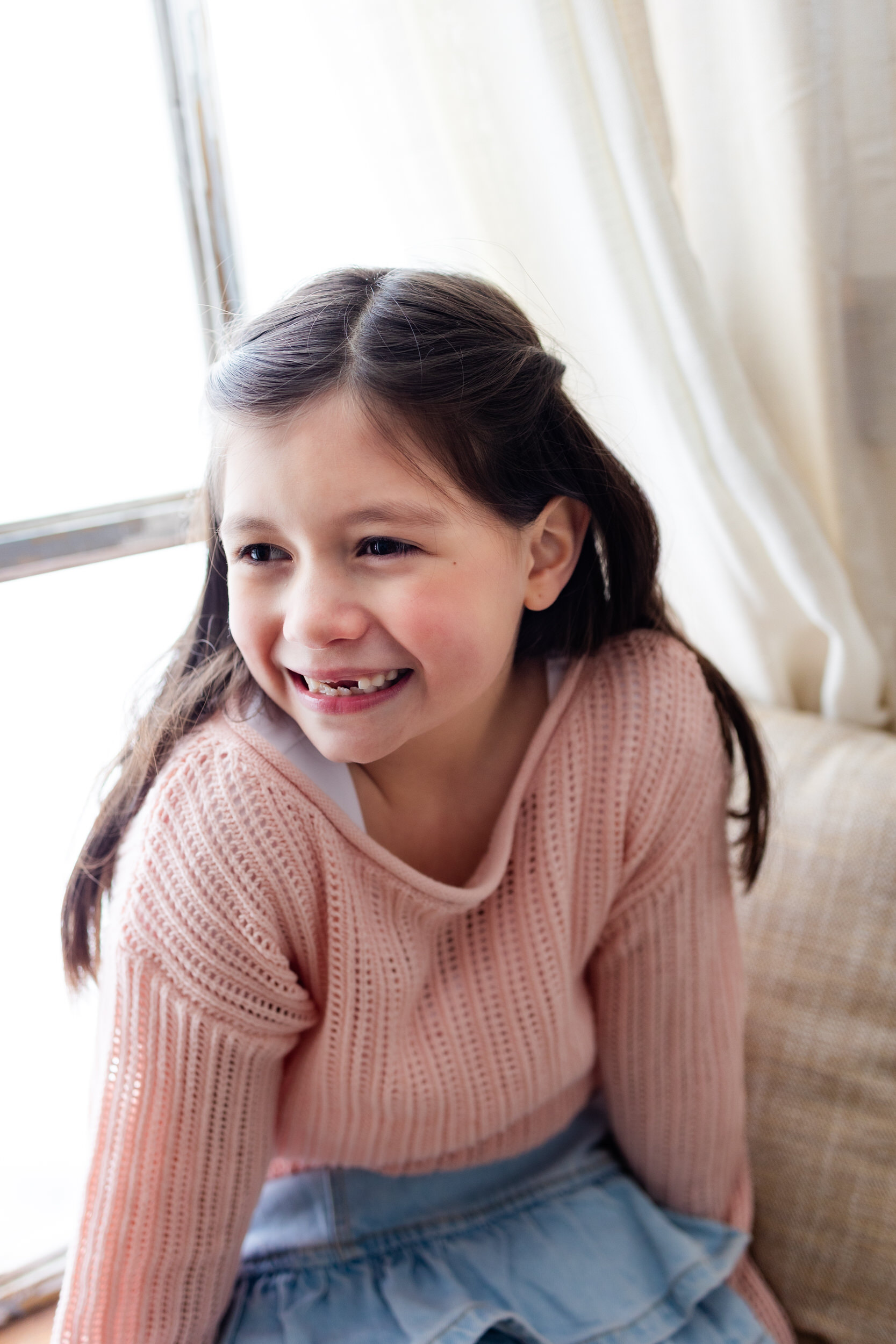 A young girl sits in a windowsill and smiles off-camera.
