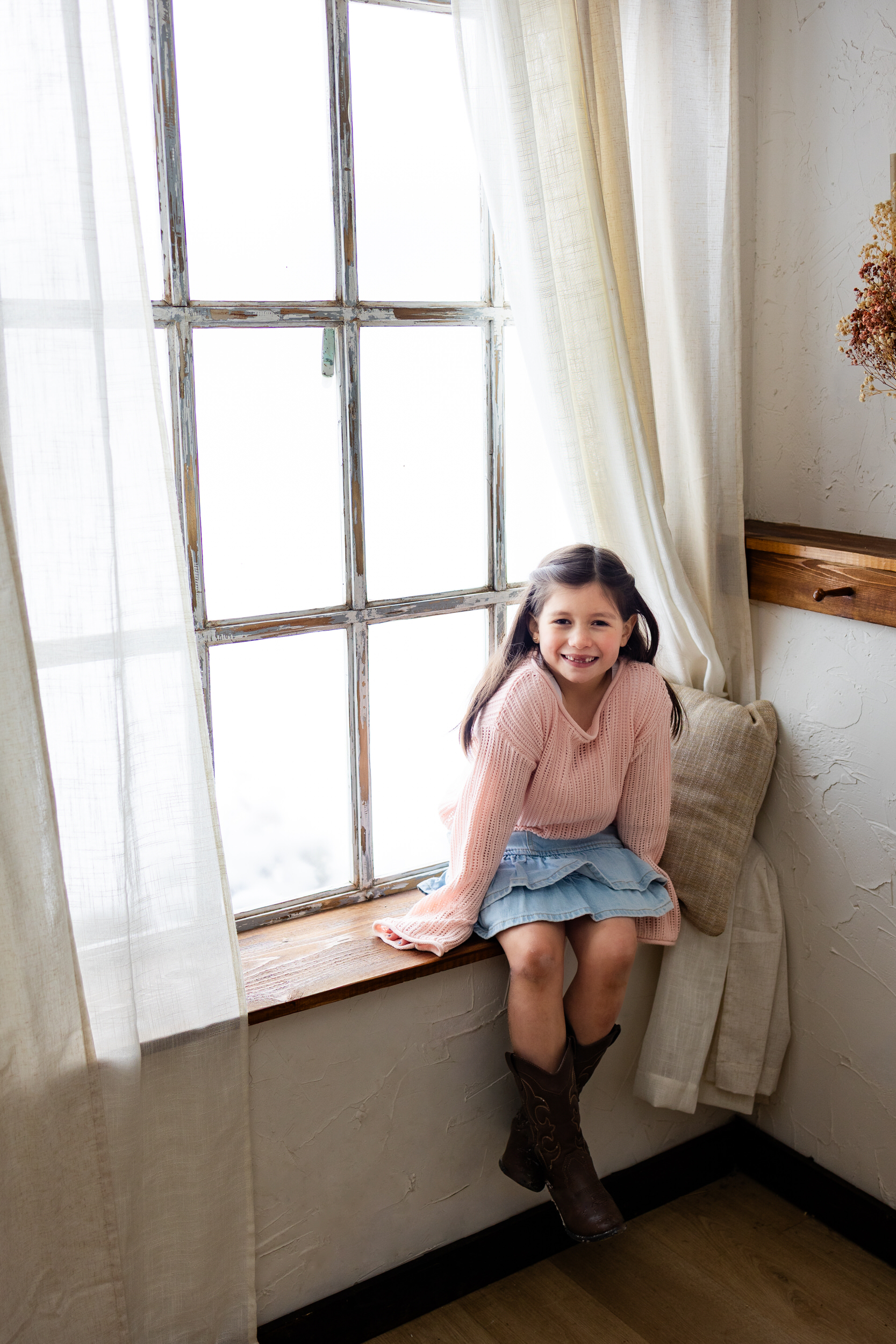 A young girl sits in a windowsill and smiles at the camera.