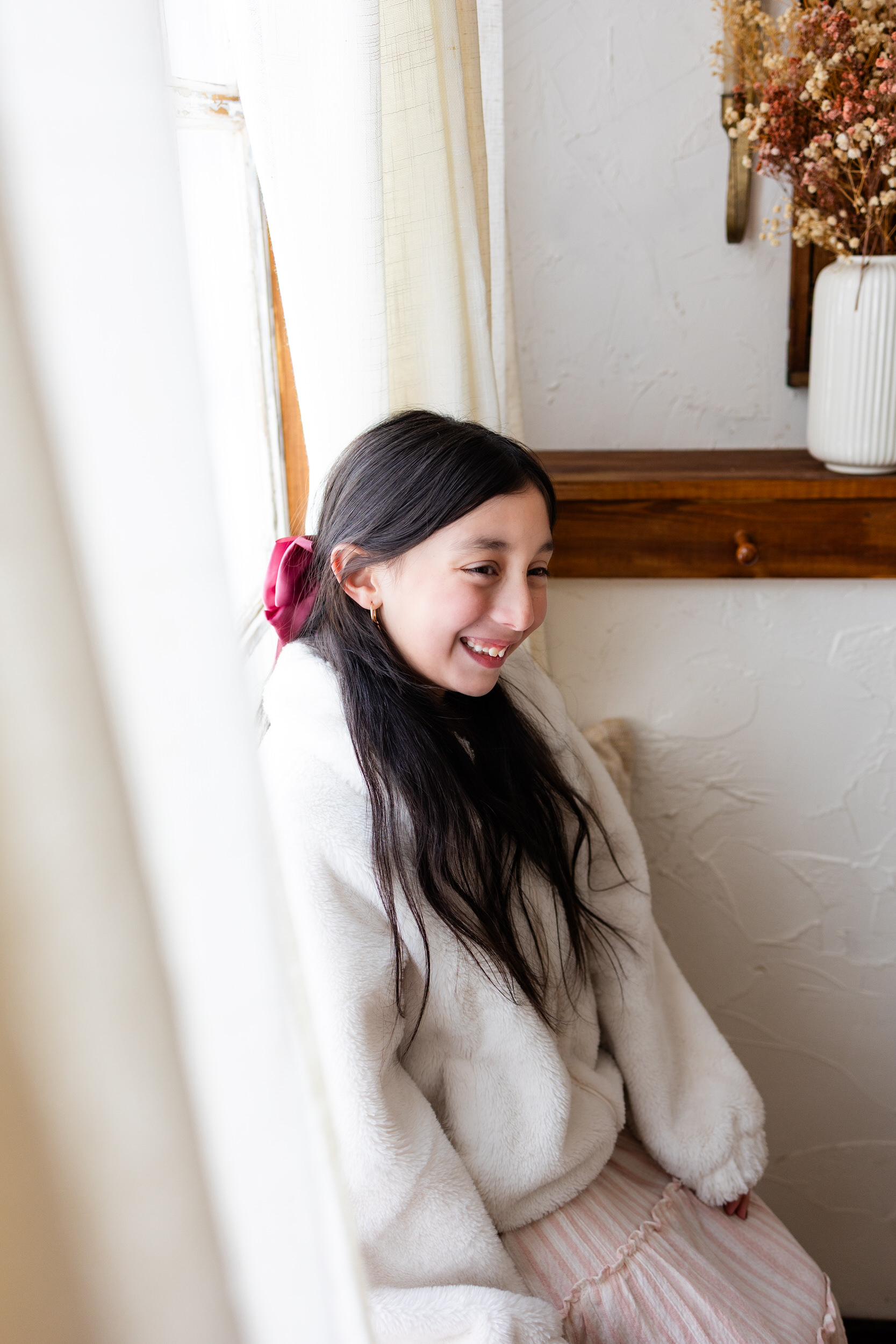 A young girl sits in a windowsill and smiles off-camera.