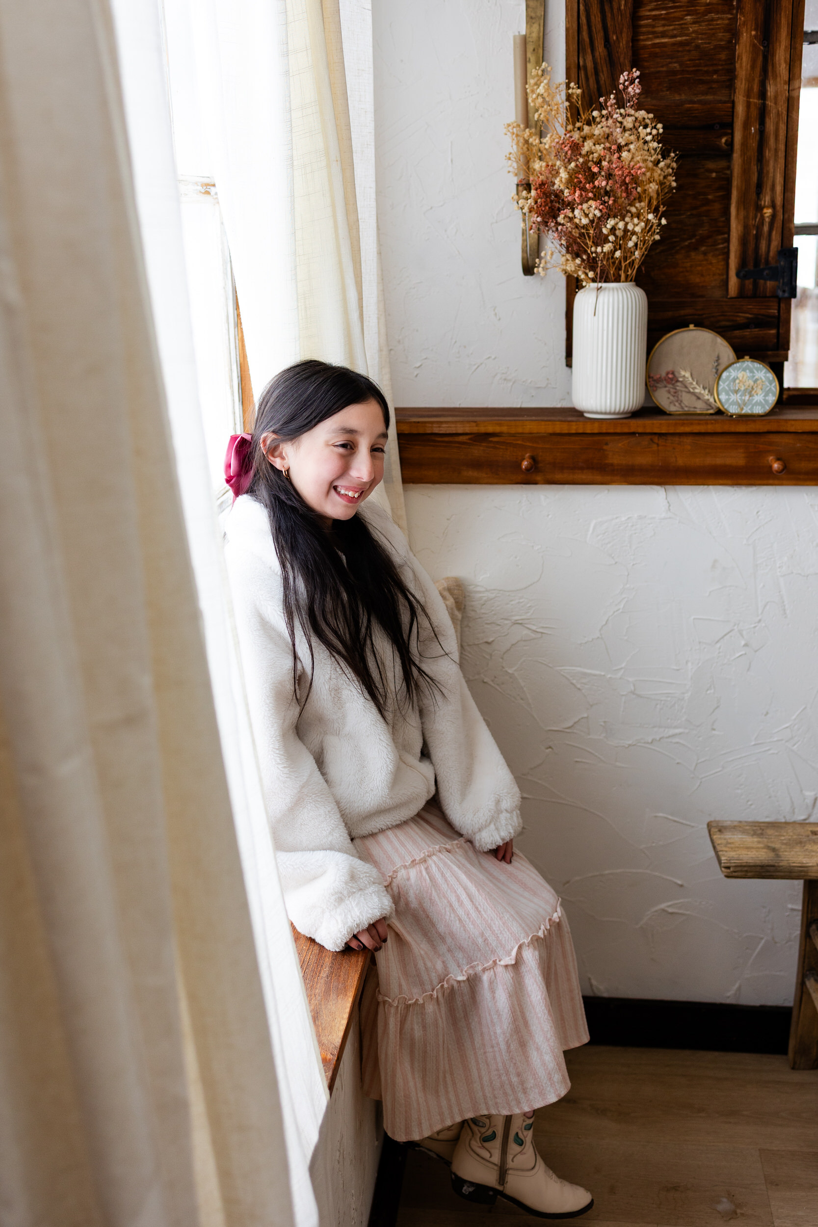 A young girl sits in a windowsill and smiles off-camera.