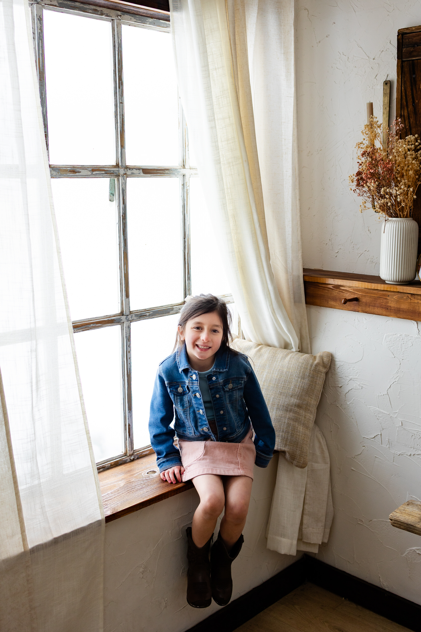 A young girl sits in a windowsill and smiles at the camera.